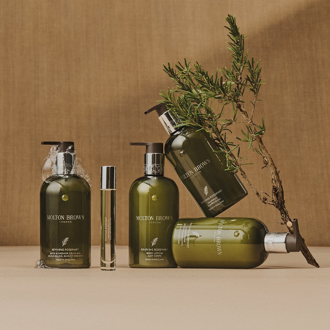 Collection of green Molton Brown skincare and bath products, including bottles of body lotion, shower gel, and a perfume, with a sprig of rosemary against a plain background.