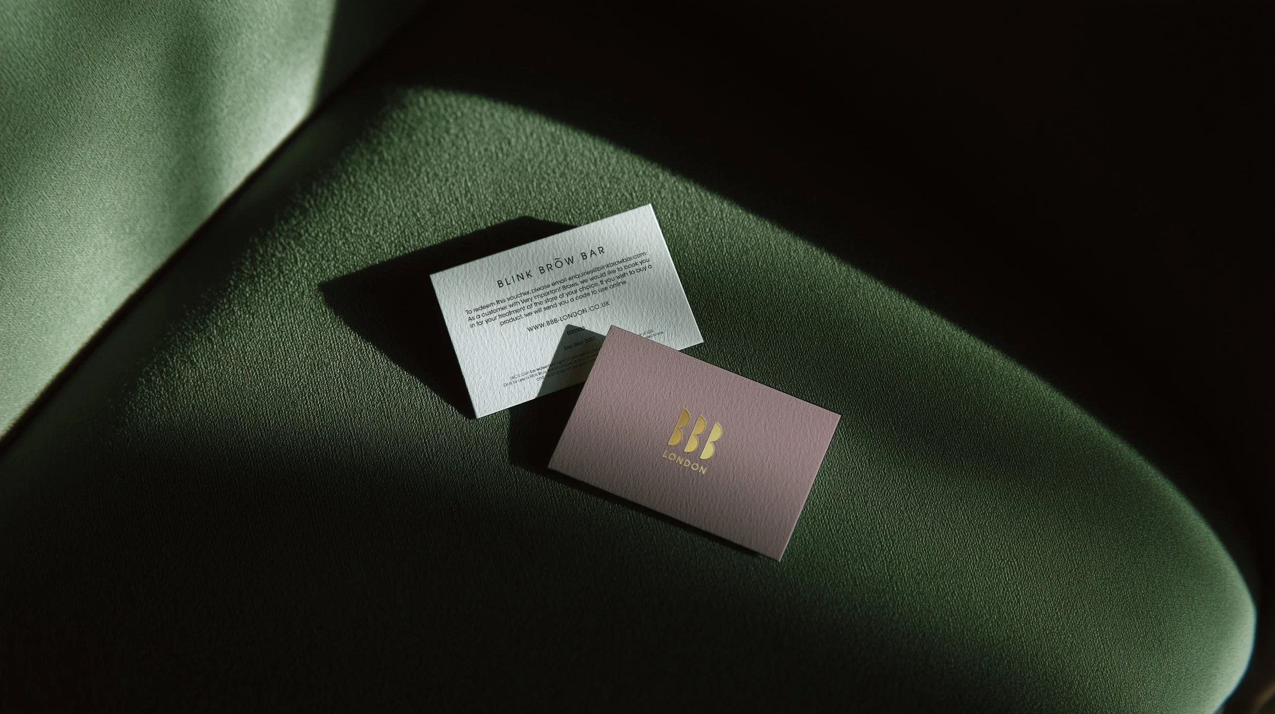 Close-up of pink card with gold BB London logo, white paper with printed text, and dark shadow on green textured surface.