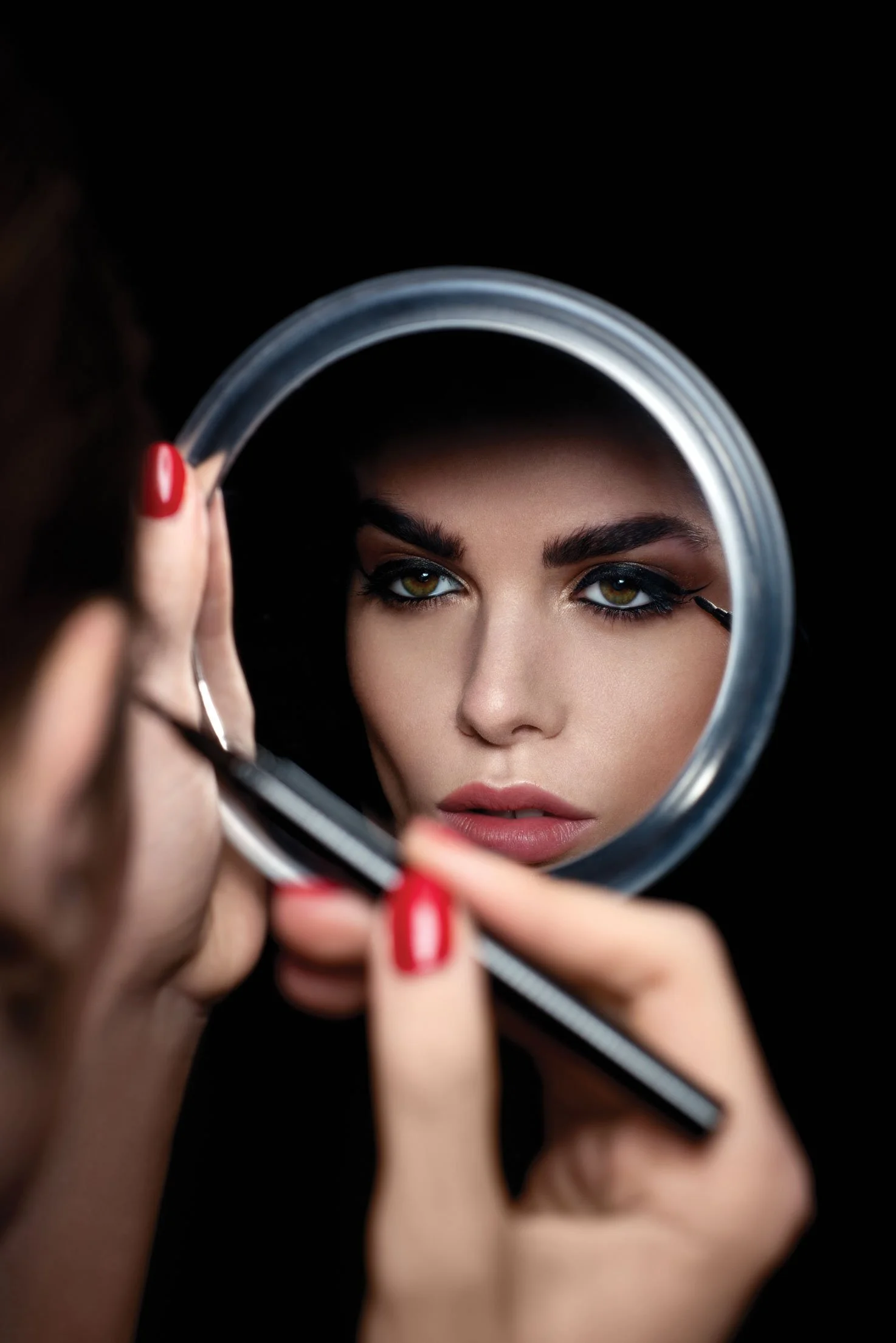 A woman with yellow-green eyes and dark makeup looking into a mirror while applying eyeliner.