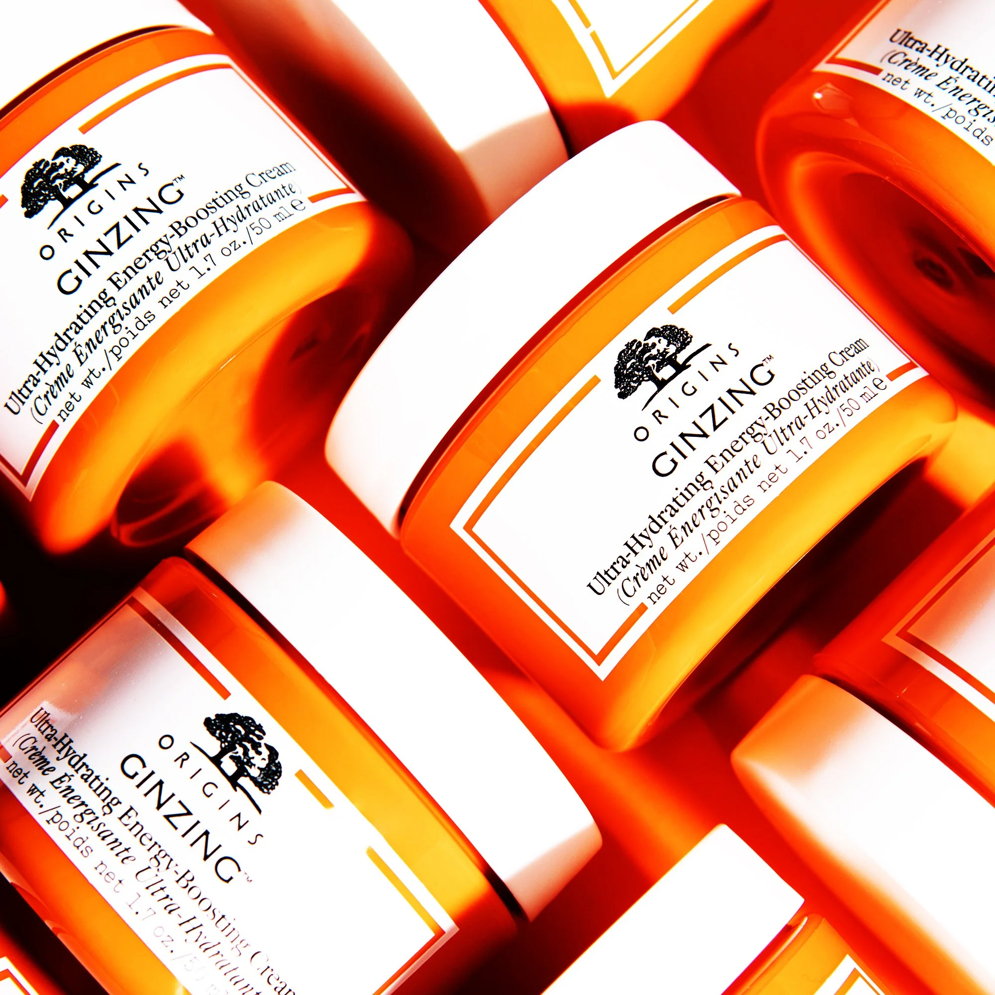 Multiple containers of Origins Ginzing Ultra-Hydrating Energy-Boosting Cream stacked together, with bright orange and white packaging and black text.