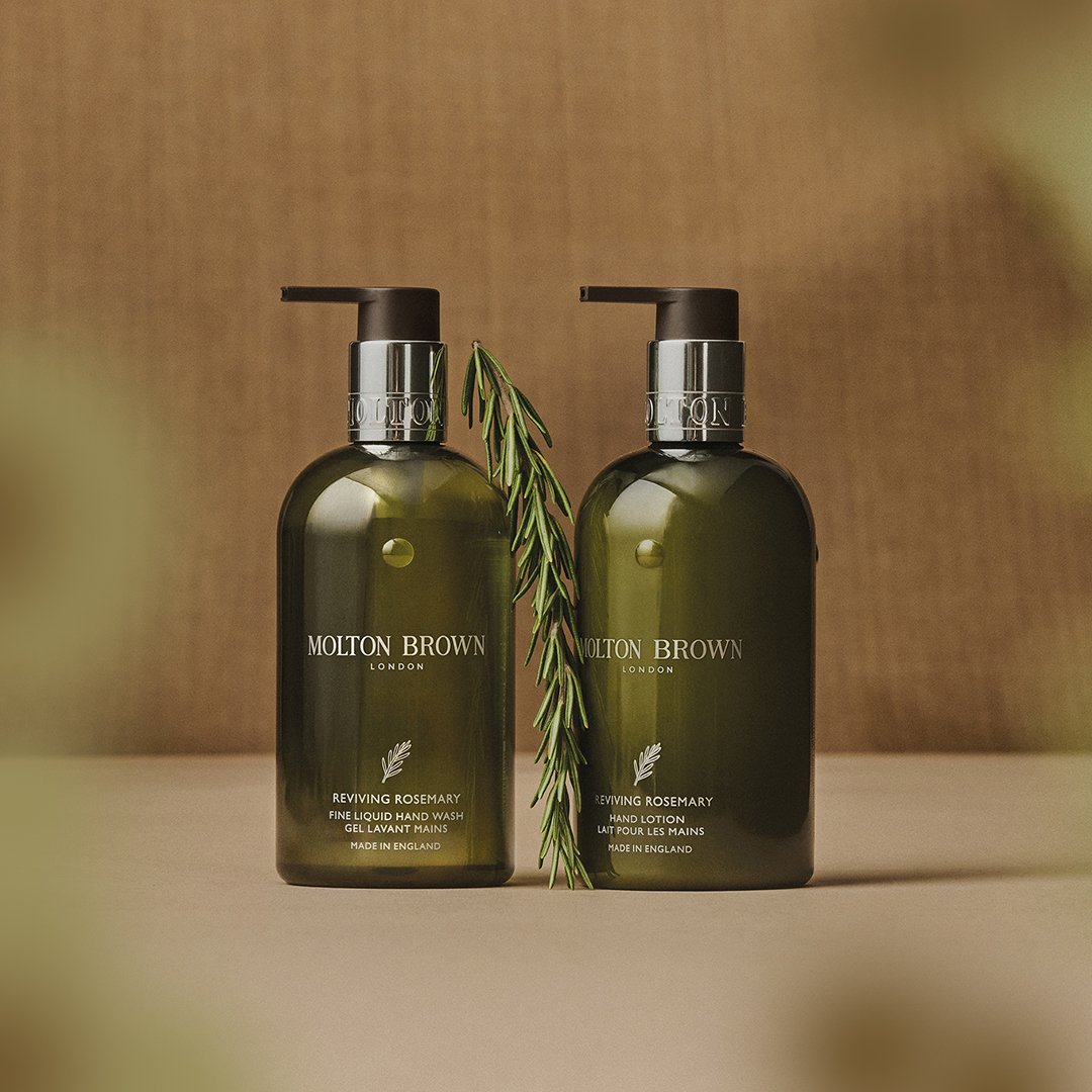 Two dark green bottles of Molton Brown hand care products with a sprig of rosemary between them, set against a warm, textured background.