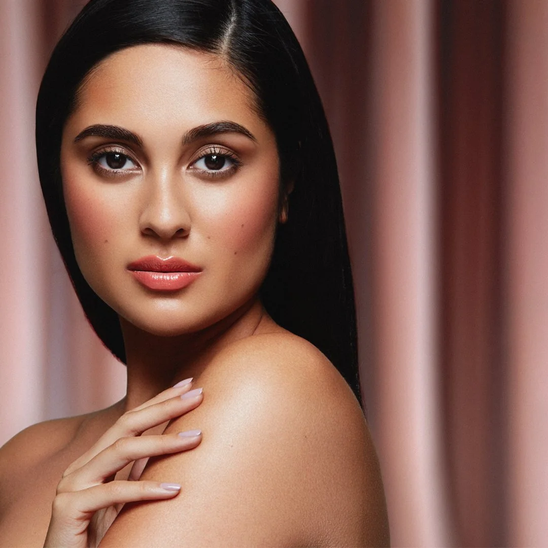 A close-up portrait of a woman with long dark hair, clear skin, light makeup, and pink lipstick, touching her shoulder with her hand, with pink curtains in the background.