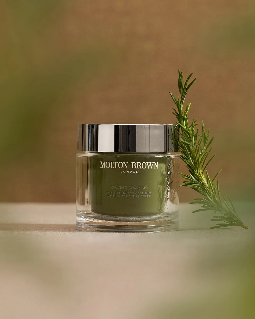 A jar of Molton Brown Reviving Rosemary exfoliating body polisher with a sprig of rosemary beside it, set on a neutral surface against a soft, blurred background.