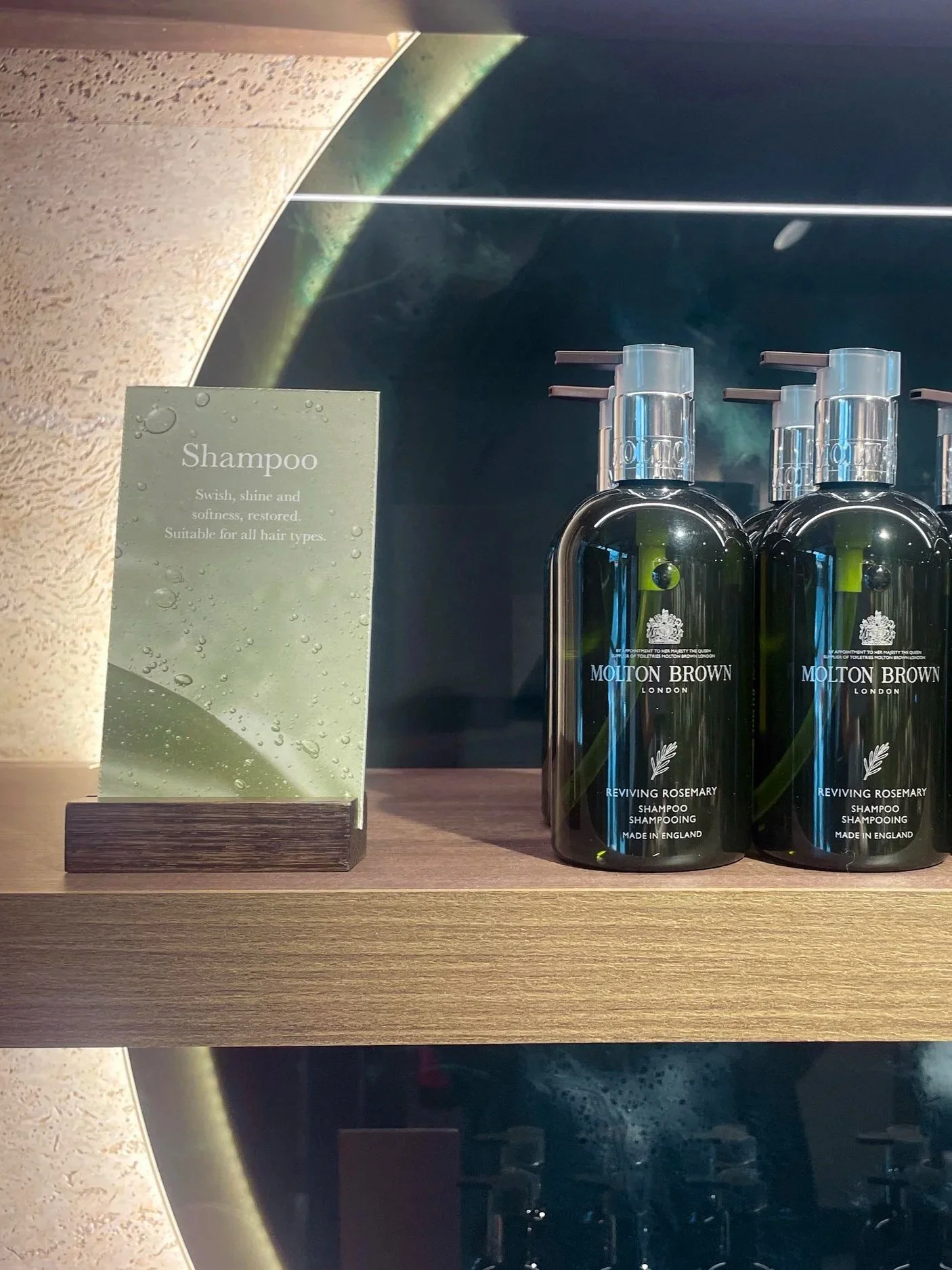 Two bottles of Molton Brown Reviving Rosemary Shampoo placed on a wooden shelf alongside a sign that reads, 'Shampoo. Swish, shine and softness, restored. Suitable for all hair types.' behind them is a mural of space, showing Earth and the stars.