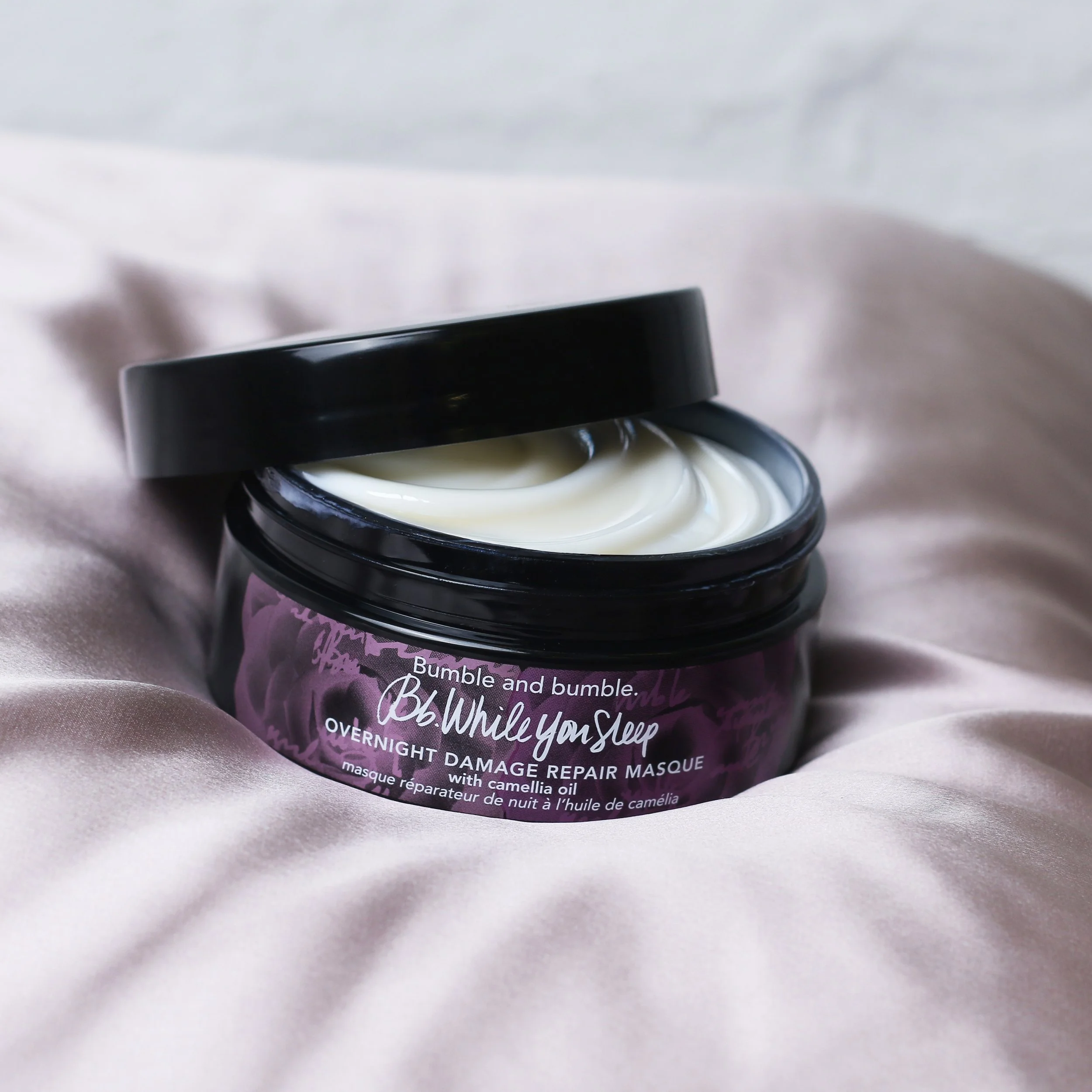 Open jar of Bumble and Bumble overnight damage repair masque with a black lid, placed on soft pink satin fabric.