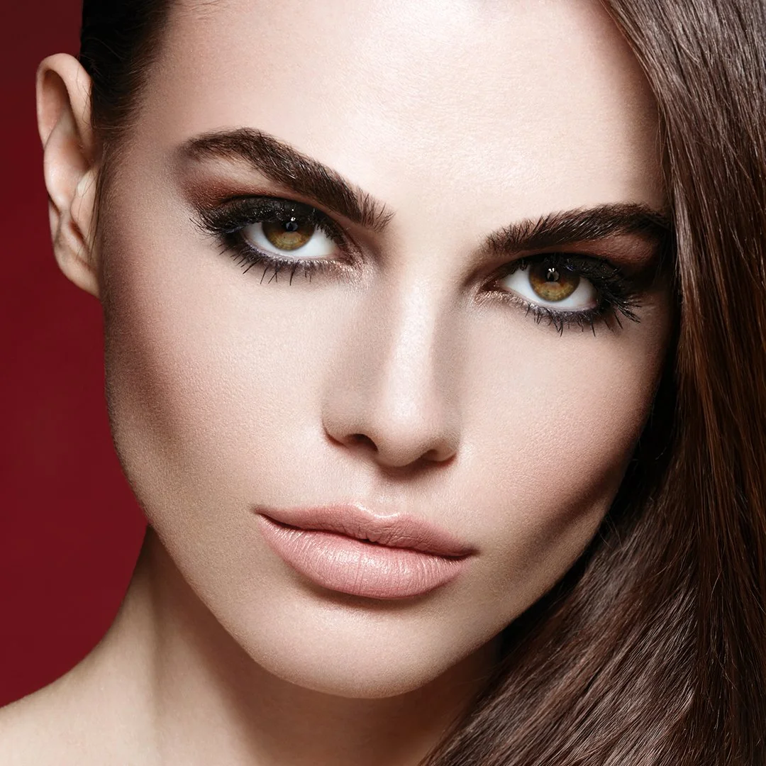 Close-up of a woman's face with makeup, including dark eye makeup, nude lipstick, and smooth skin, against a red background.