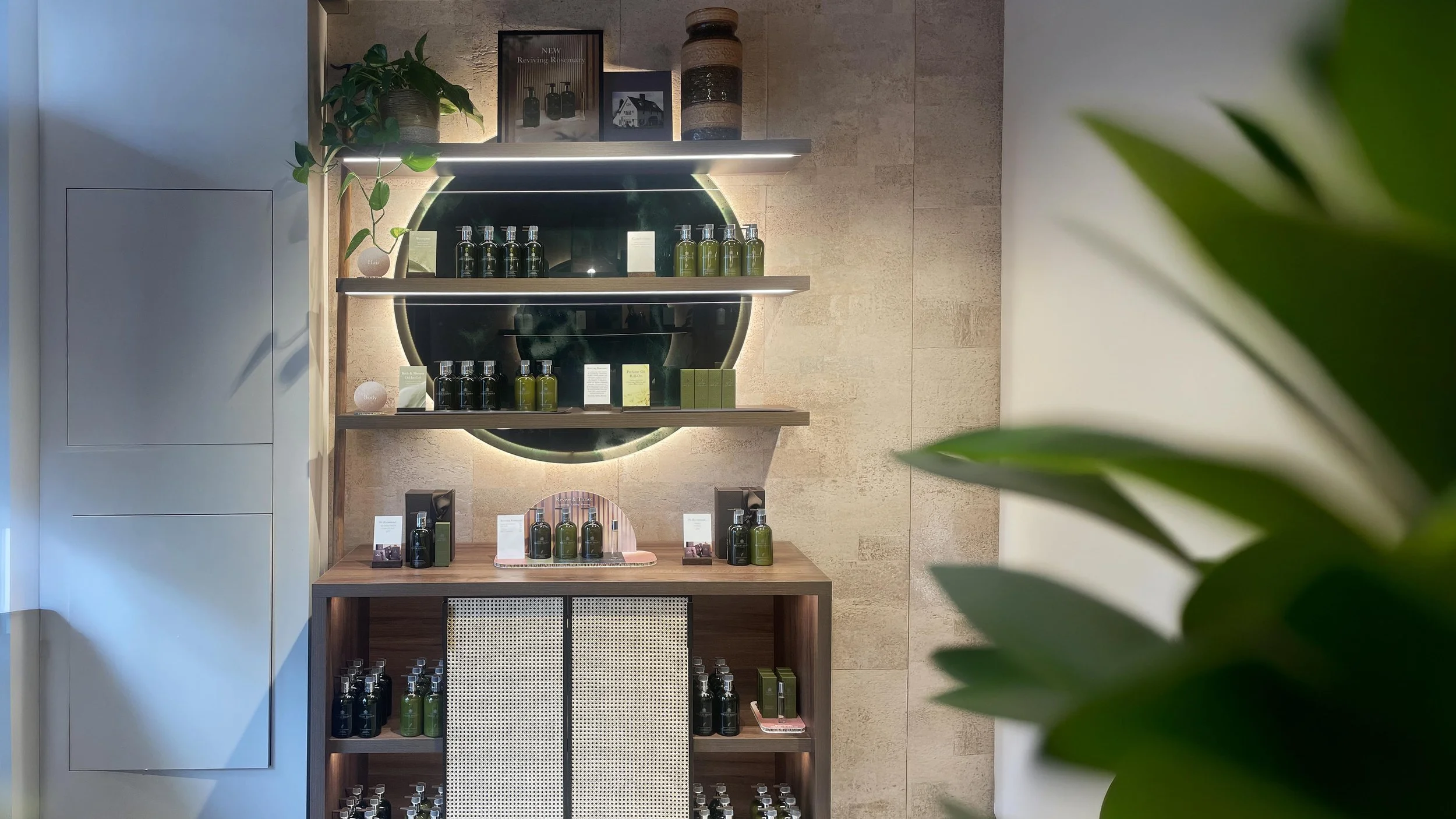 Display shelf with green and black bottles of body and skincare products, a small plant, framed pictures, and decorative jars against a beige tiled wall in a retail or spa setting.