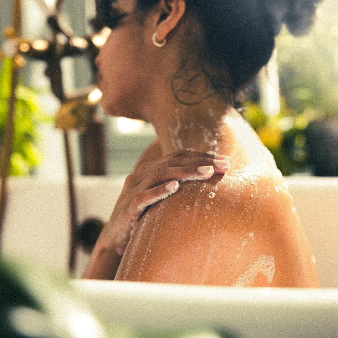 A woman taking a bath, with soap bubbles on her shoulder and neck, in a bathroom with natural light and greenery in the background.
