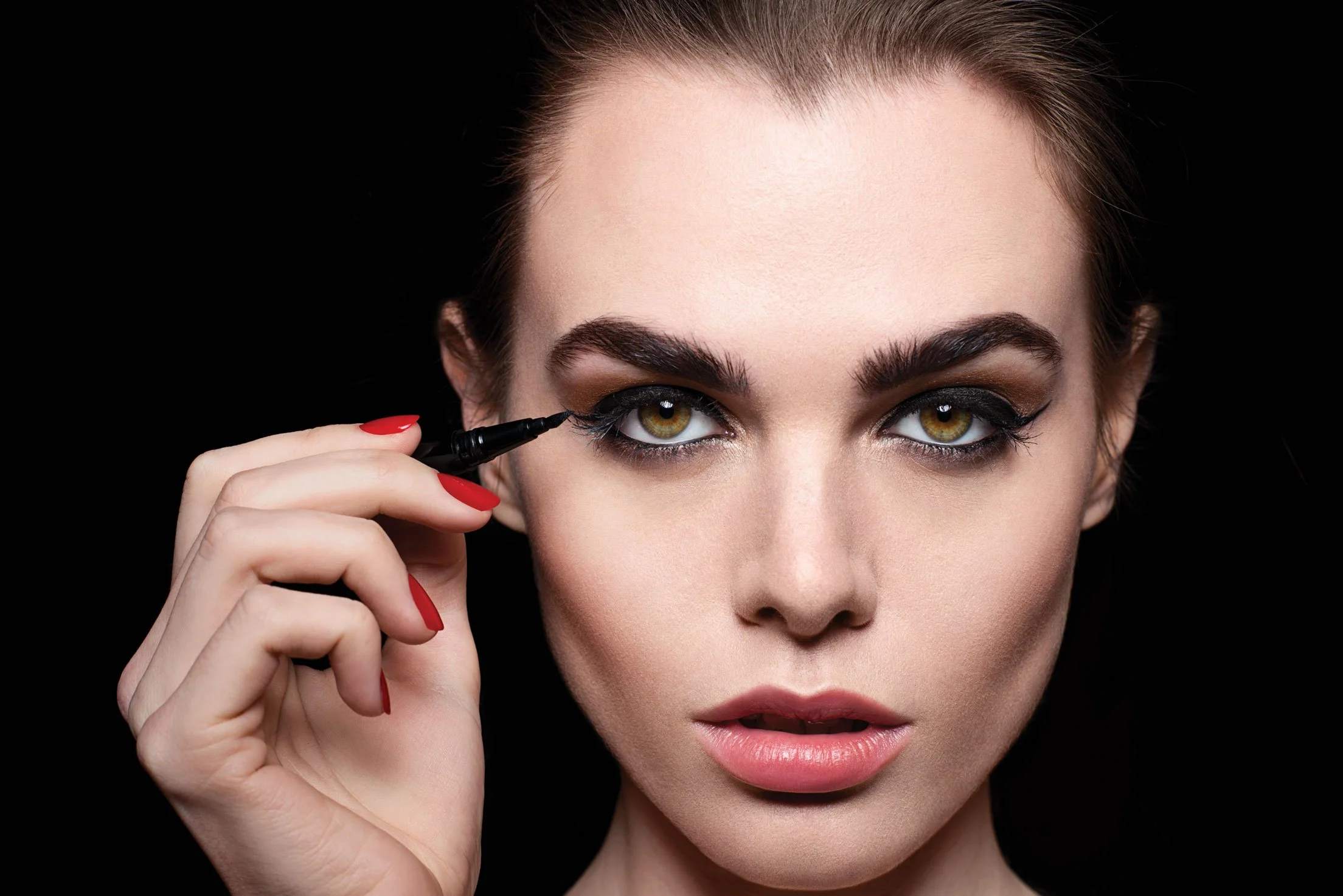 Close-up of a woman applying black eyeliner to her eye against a black background.