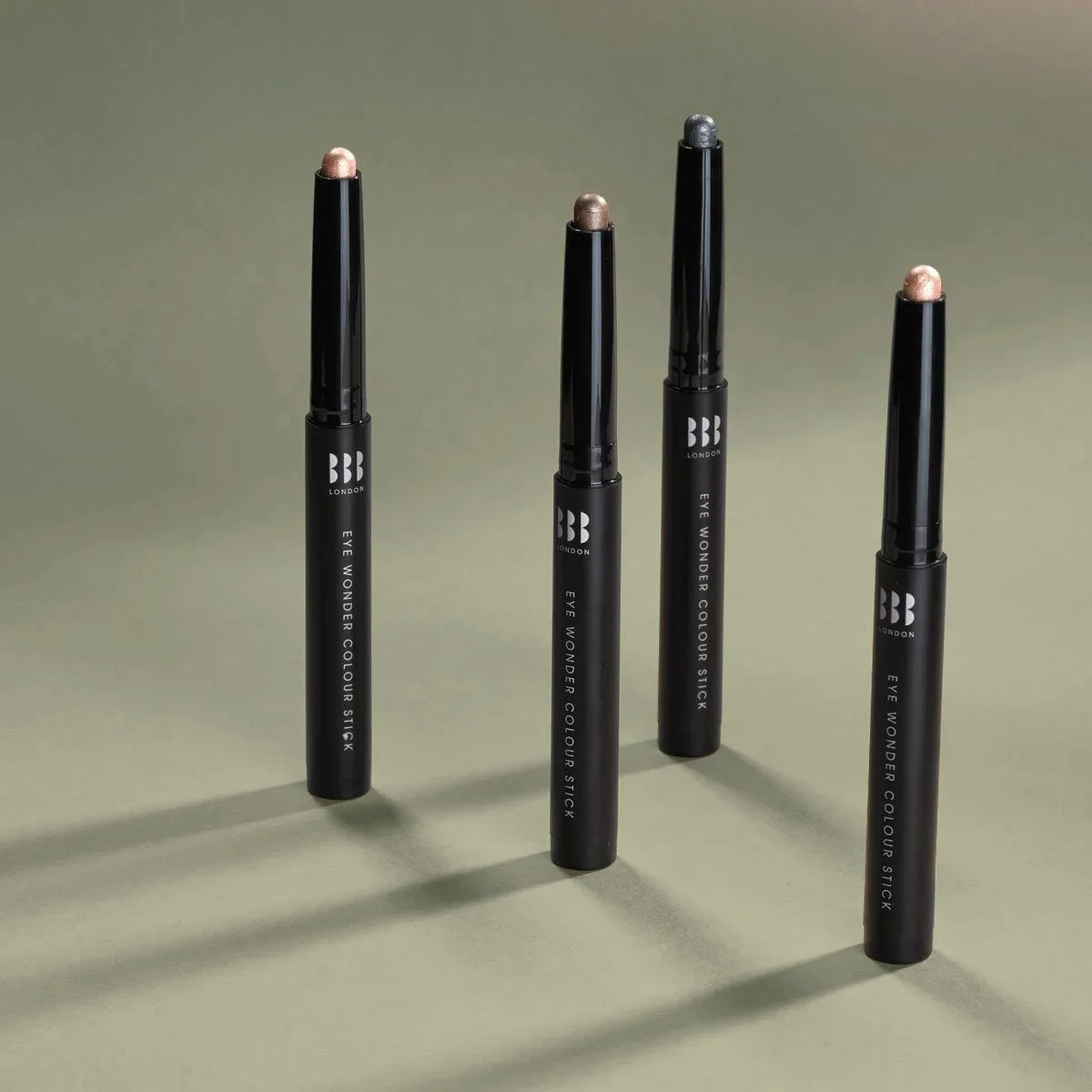 Four black eye shadow sticks with different shades, standing upright on a light green surface, casting shadows.