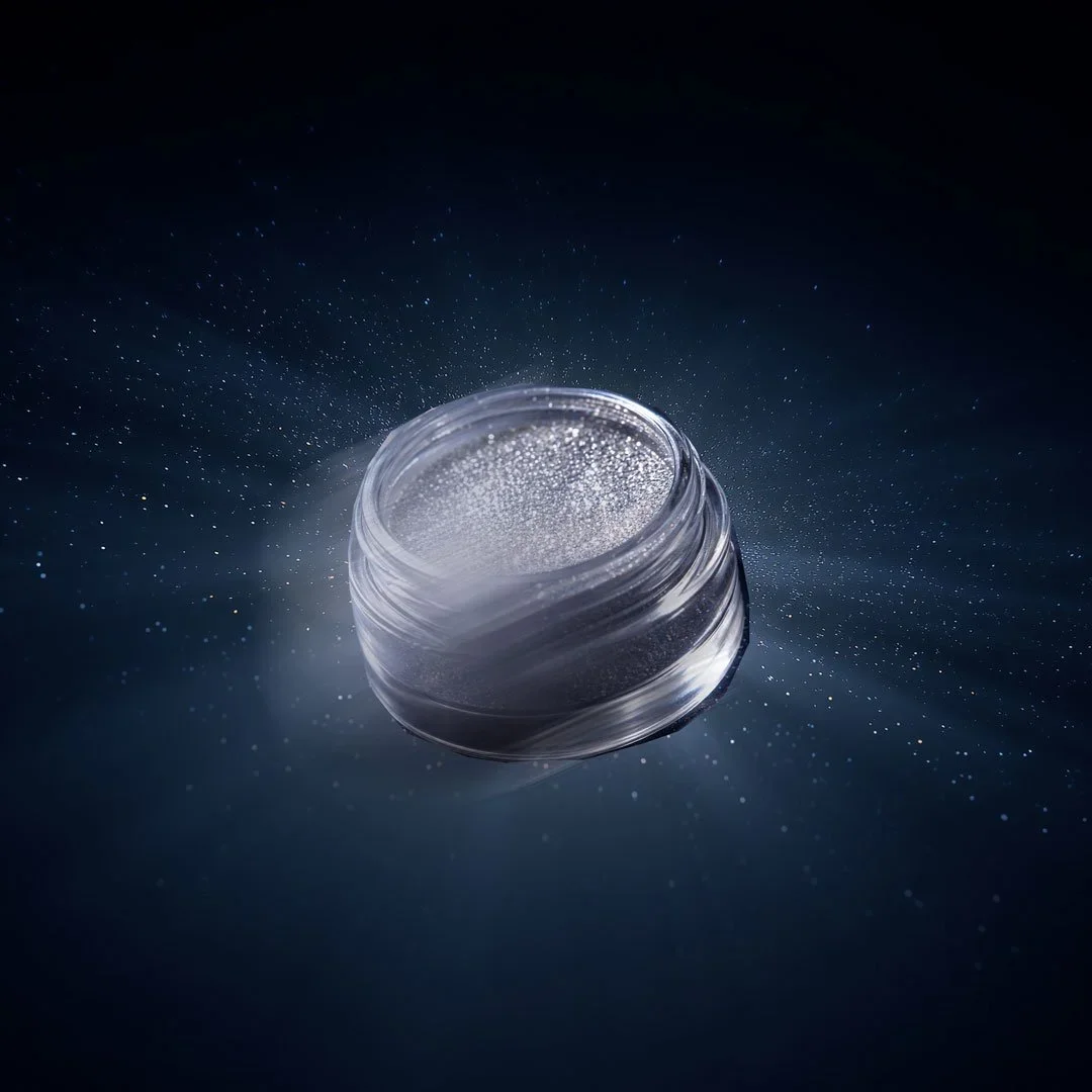 Open jar of loose glitter or cosmetic shimmer with scattered particles surrounding it on a dark background.