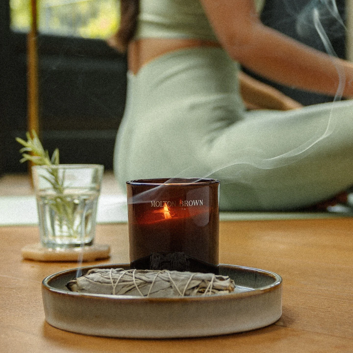 A tray with smudge sticks and a candle on a wooden table, with a person in the background wearing light-colored pants and a striped shirt.