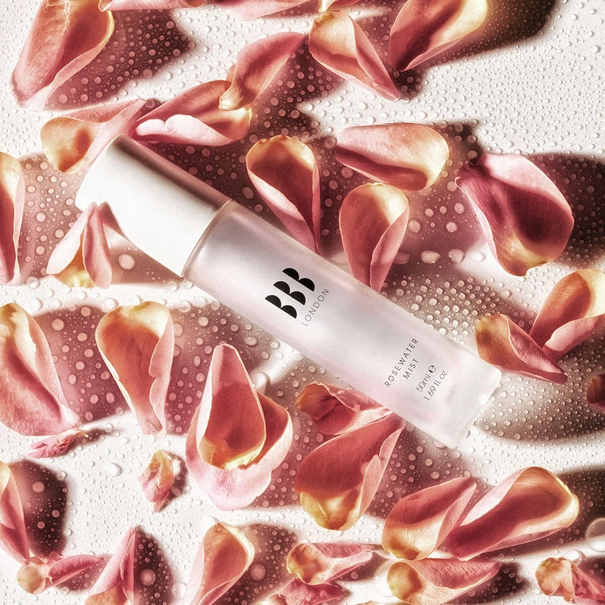 A white bottle of BB London Rosewater Mist lying among scattered pink and peach rose petals with water droplets on a textured surface.