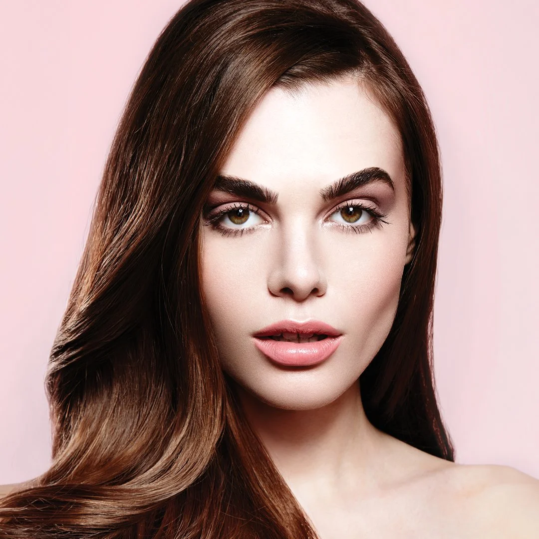 Close-up of a woman with long, wavy brown hair and light makeup against a pink background.
