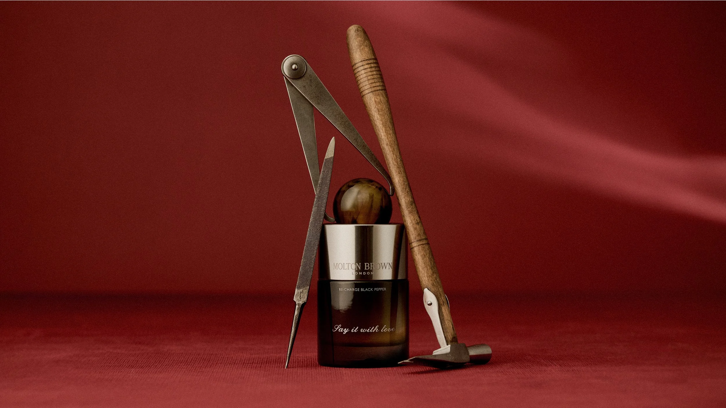 Layered Molton Brown product still life with sculptural props and rich red textured backdrop introducing the Evergreen brand world.
