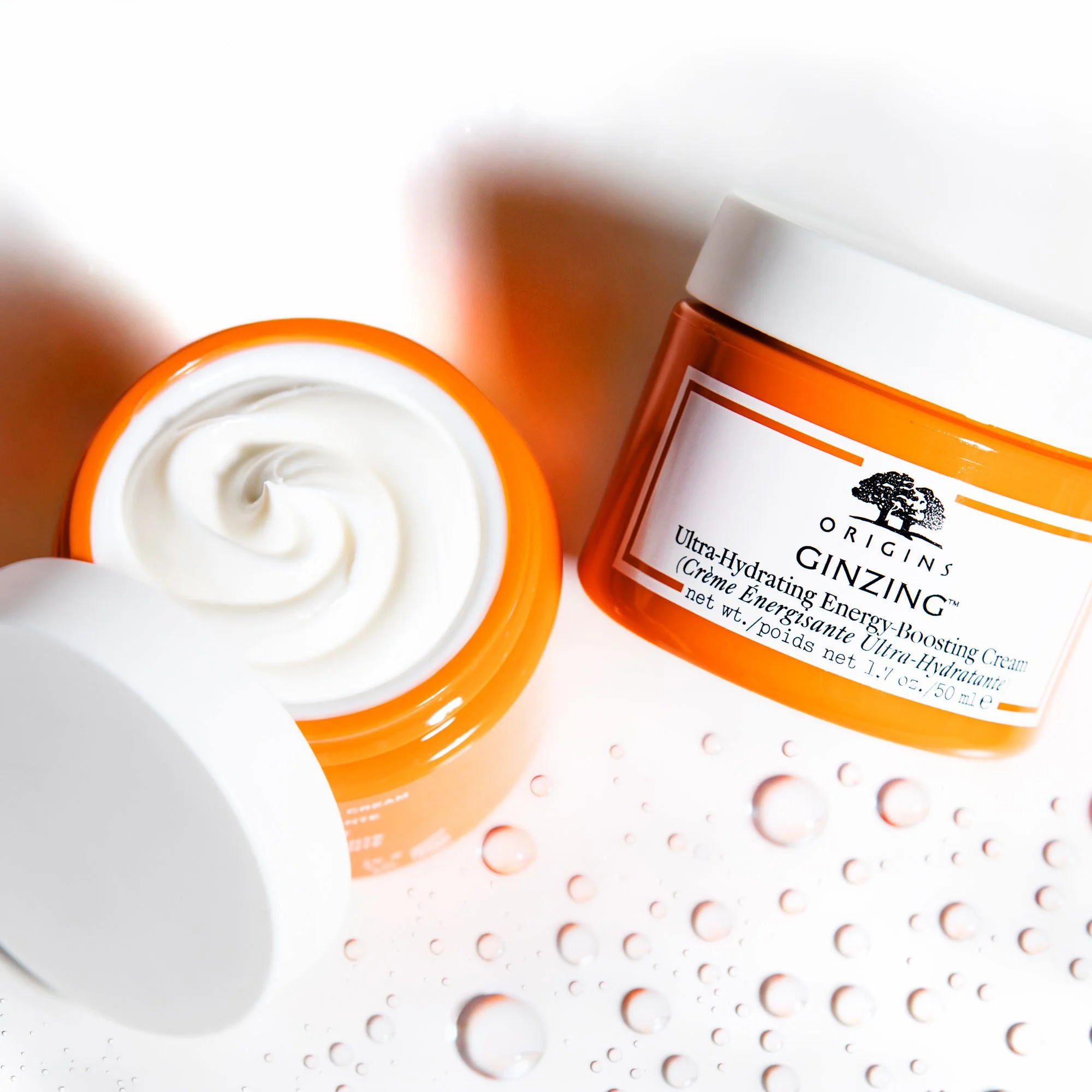 Open jar of Ginzing Ultra-Hydrating Energy-Boosting Cream with white cream visible inside, placed on a white surface with water droplets around.