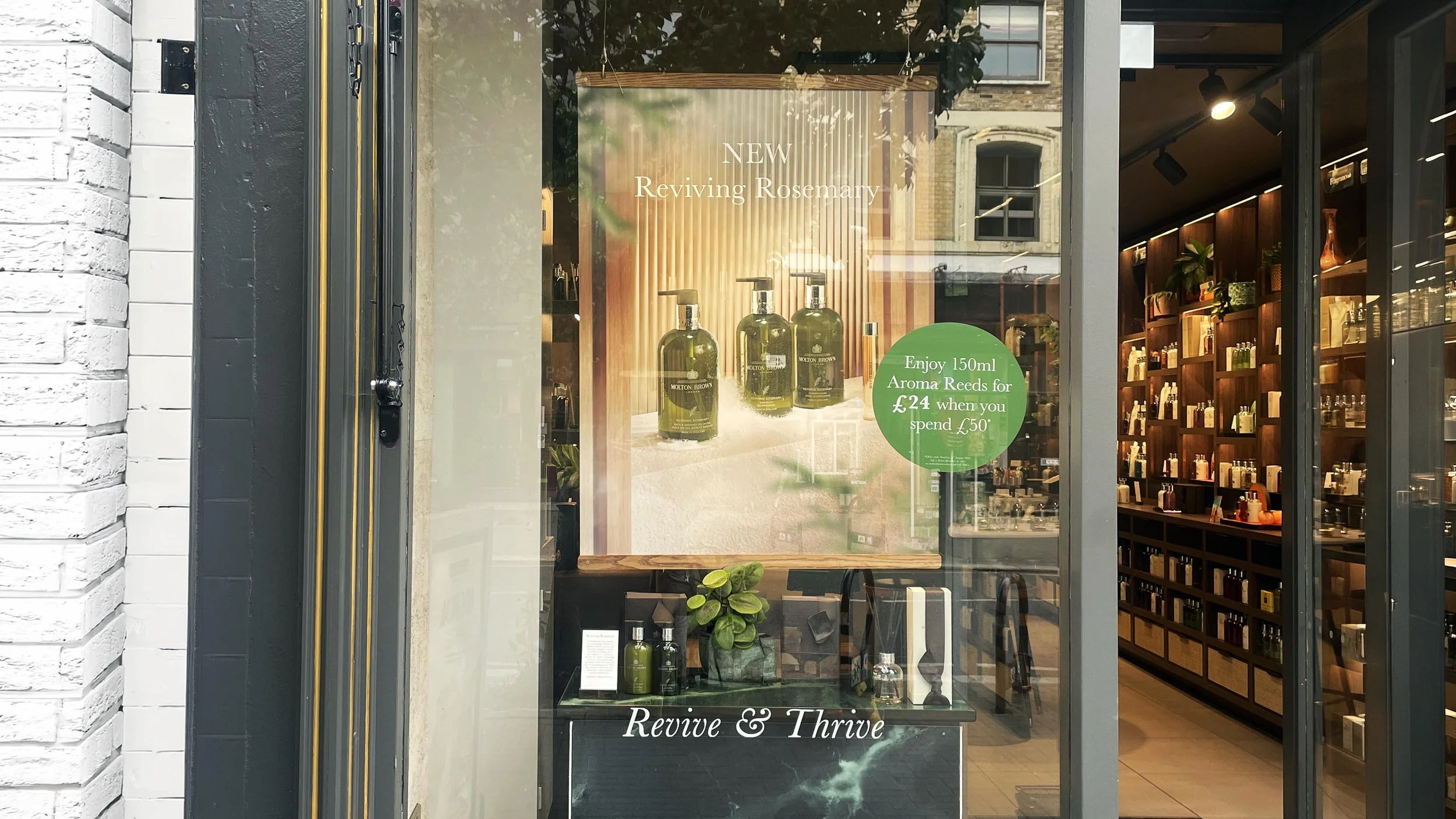 Storefront window displaying bottles of aromatherapy oil with text promoting a new product called Reviving Rosemary and a special offer for aroma reed diffusers. Inside the store, shelves with various bottles and products are visible.