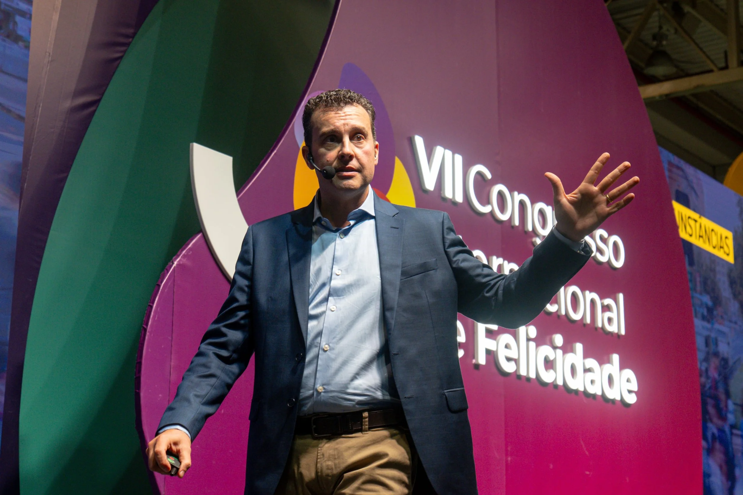 Homem de terno azul escuro, camisa azul clara, com microfone na cabeça, fazendo uma apresentação em um palco com fundo roxo e texto em português.