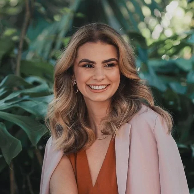 Mulher com cabelo cacheado e sorriso, ao ar livre, com árvore ao fundo.