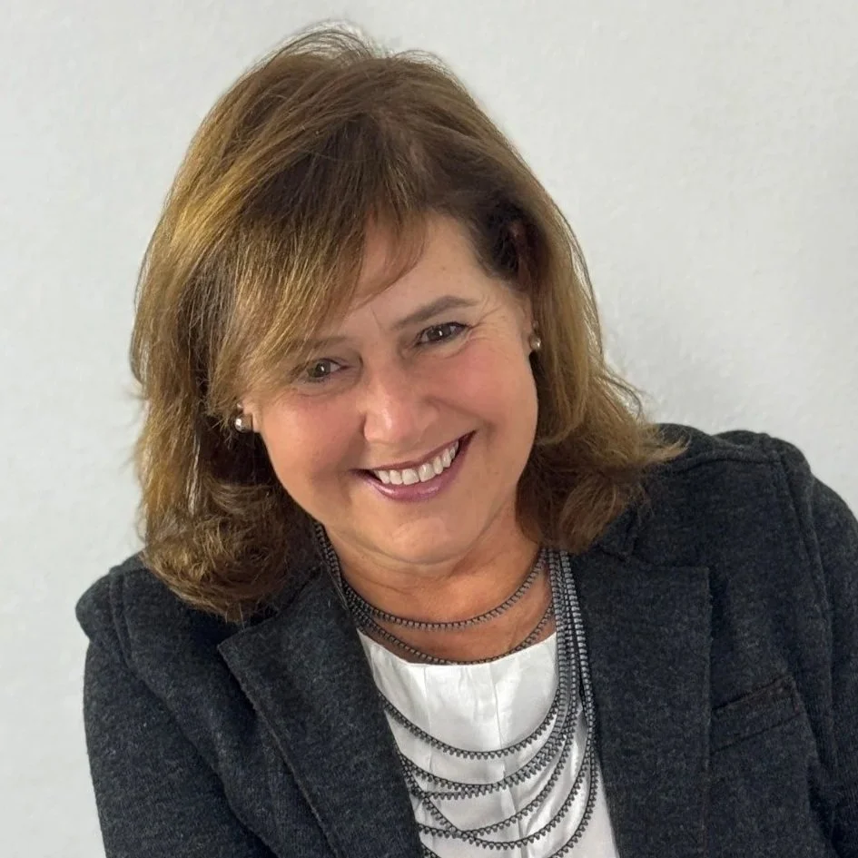 Mulher com cabelo castanho curto, sorrindo, usando suéter branco, fundo cinza