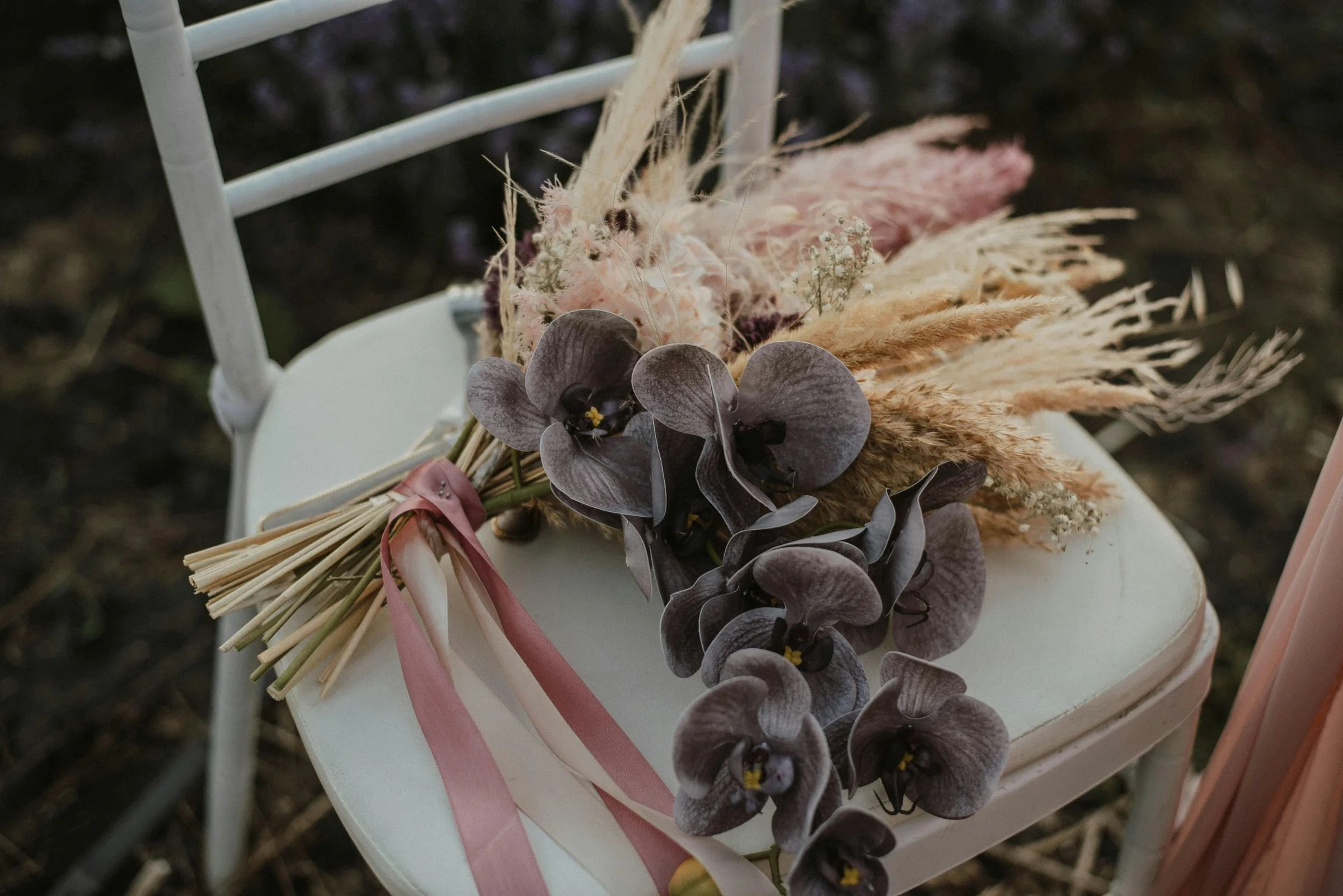 A bouquet of purple orchids, dried grasses, and pink ribbons rests on a white chair—perfect for wedding business branding.