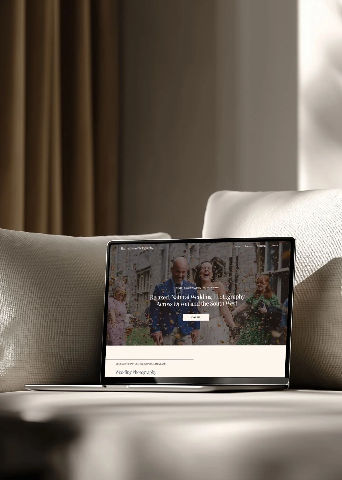A tablet displaying a wedding photography website with a happy bride and groom walking outside of a church, surrounded by guests celebrating, placed on a white surface between two beige pillows, with beige curtains and a window in the background.