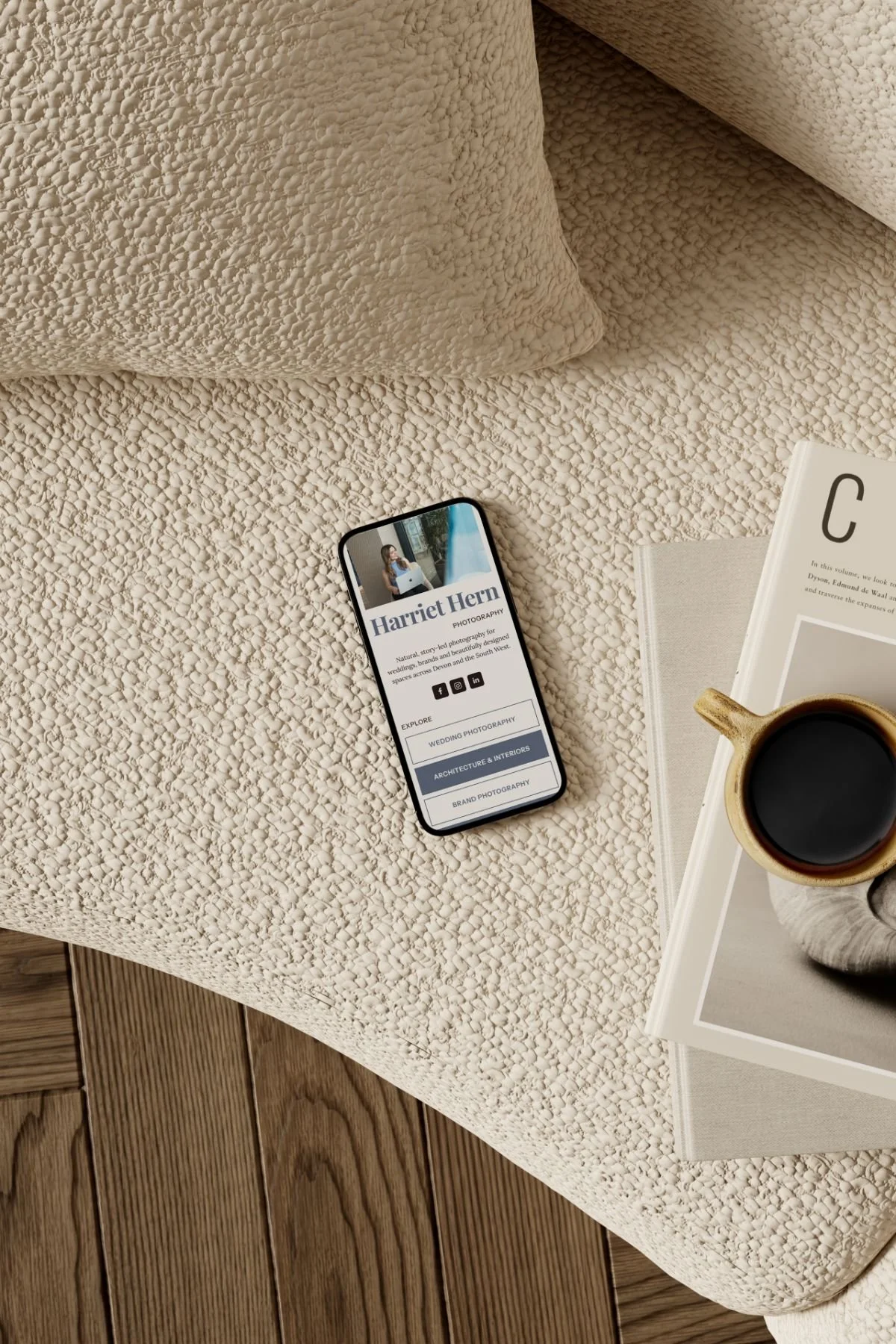 A smartphone displaying a photographer’s website, a cup of black coffee on top of a magazine, and a book on a textured beige bedspread with hardwood floors in the background.