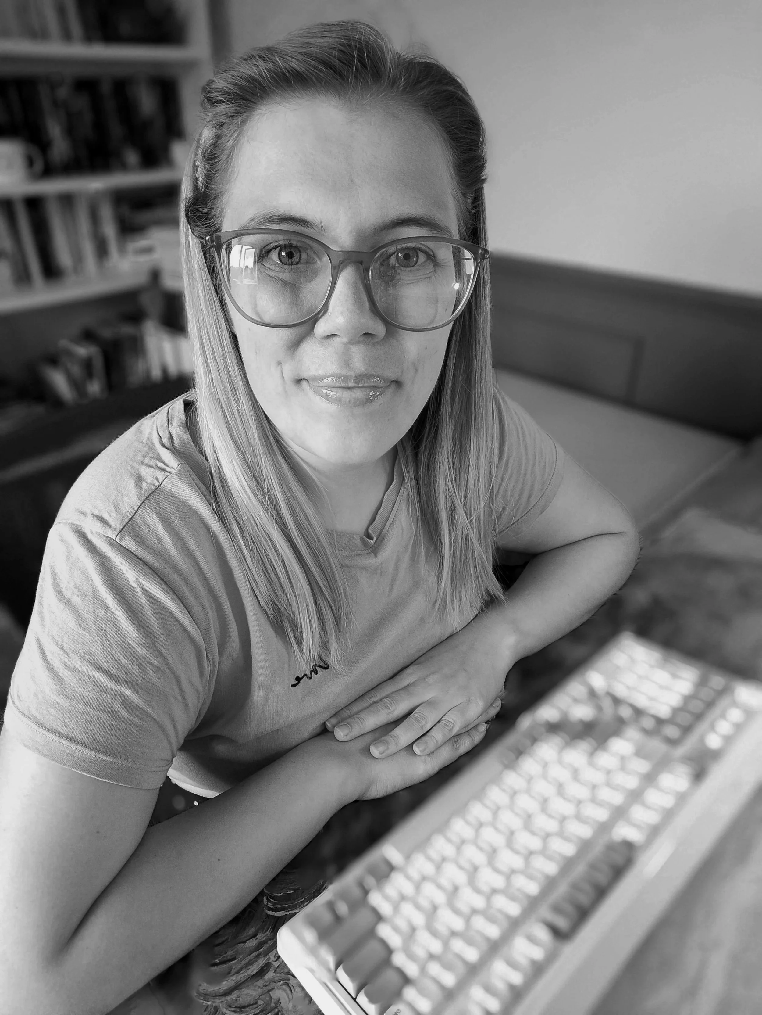 A woman with glasses and long hair sitting at a desk with a computer keyboard in front of her, in a room with bookshelves in the background.
