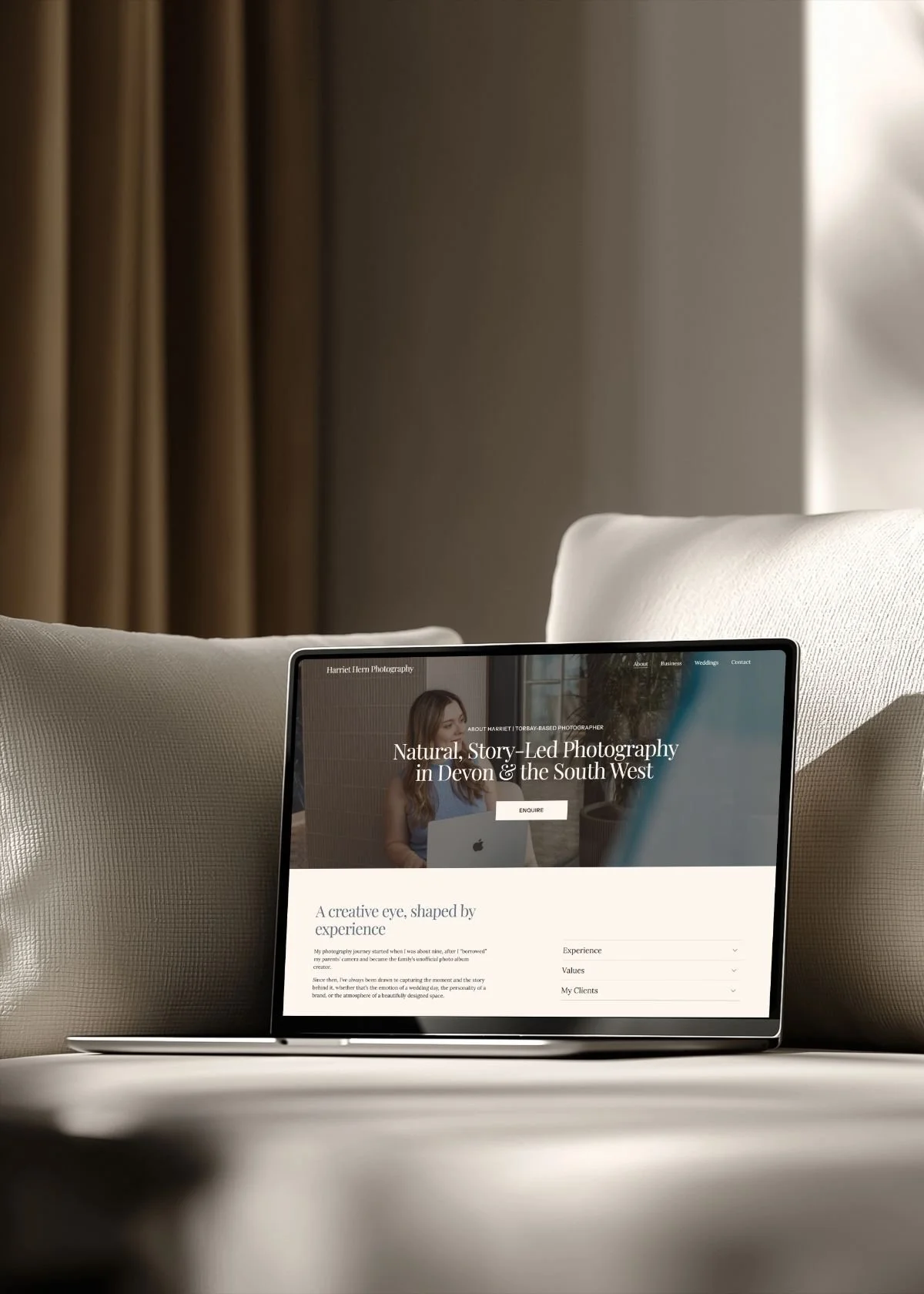 A tablet displaying a photography website on a white surface, with beige cushions on either side and beige curtains in the background.