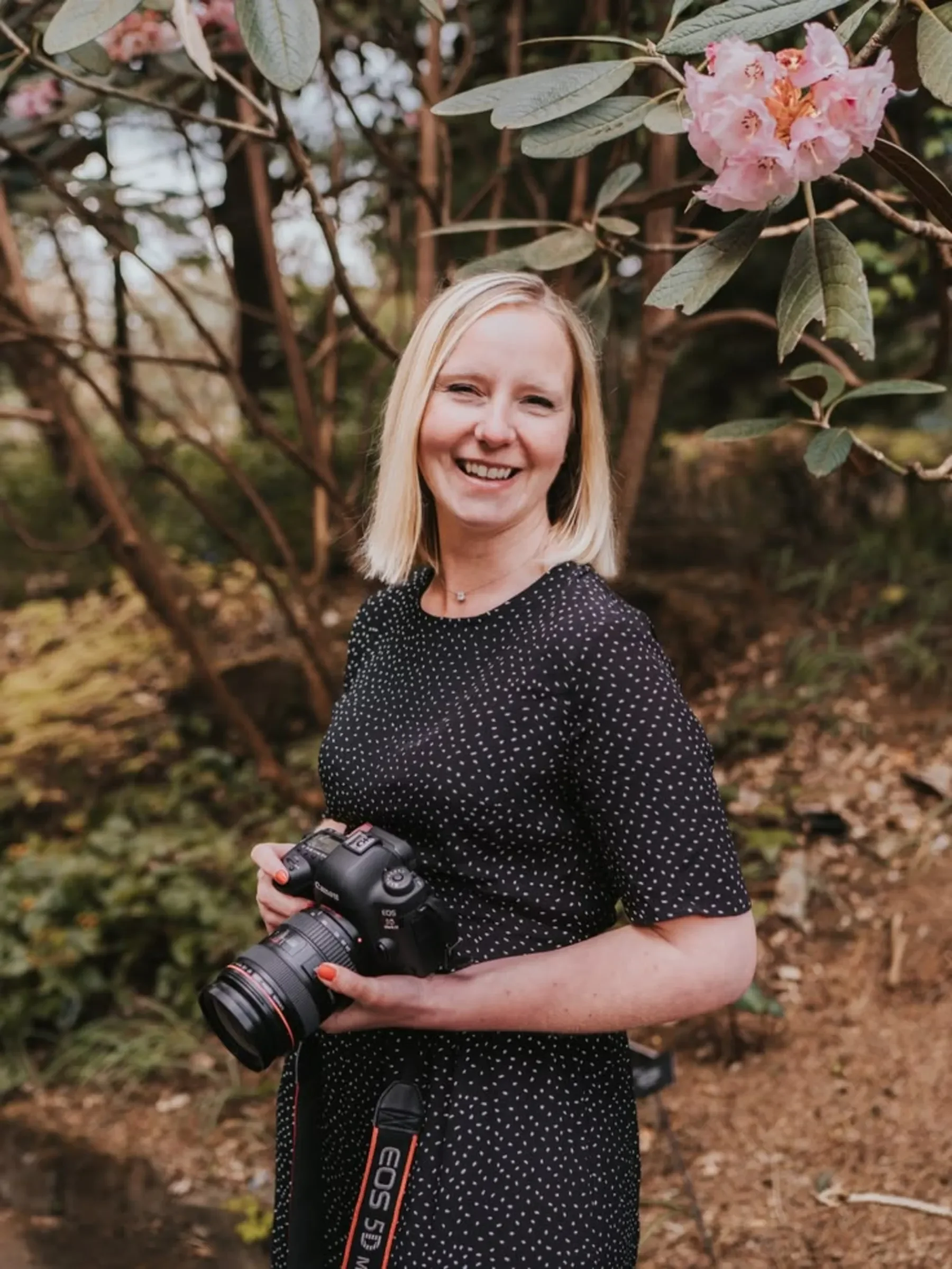 Smiling woman with a camera stands outdoors among trees and pink flowers, perfect for wedding photographer branding.