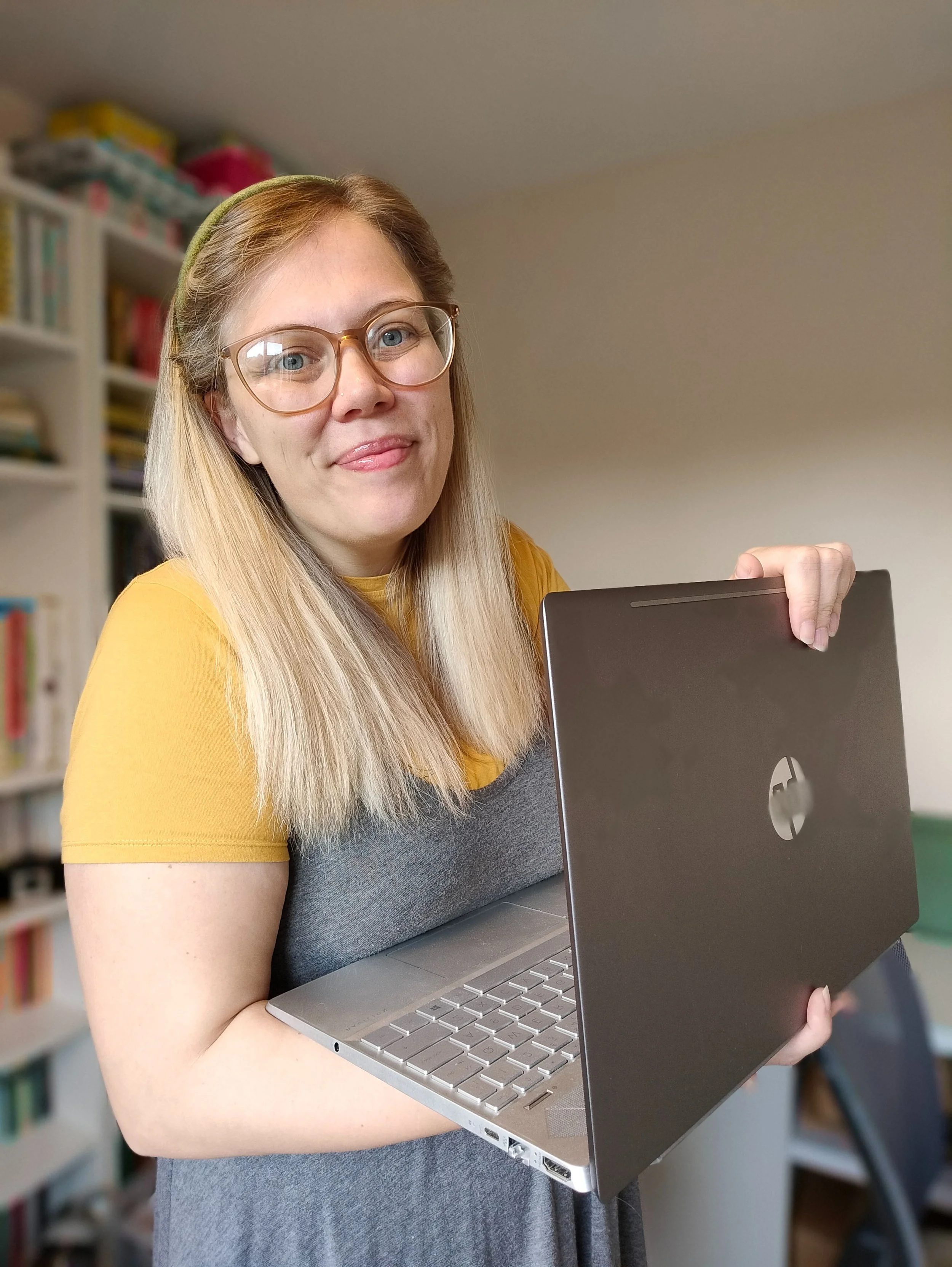 Woman with long blonde hair, glasses, and a yellow and gray shirt holding an open laptop in a room with bookshelves.