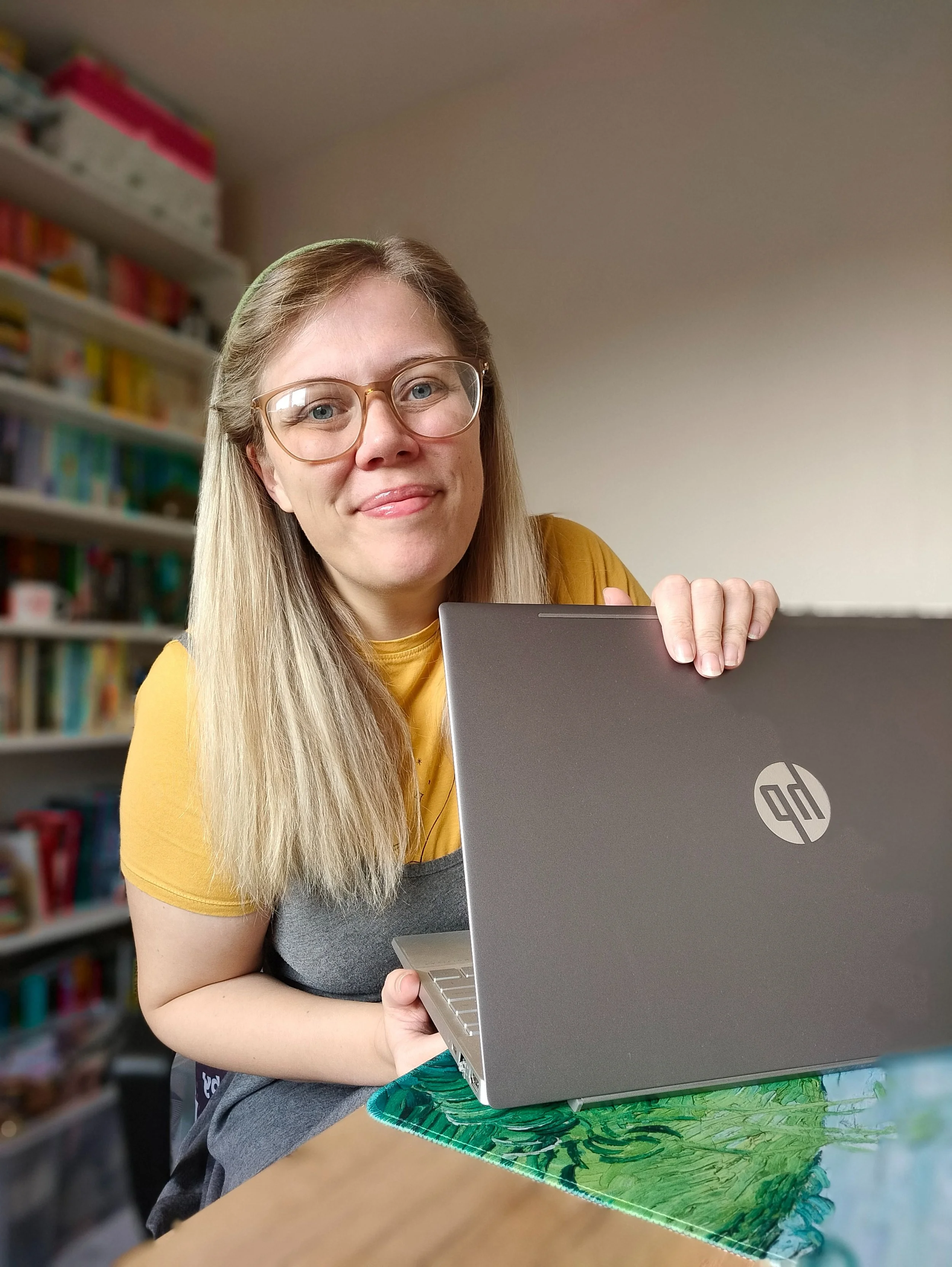 Woman with glasses and blonde hair sitting at a table with an HP laptop, smiling at the camera, with bookshelves in the background.