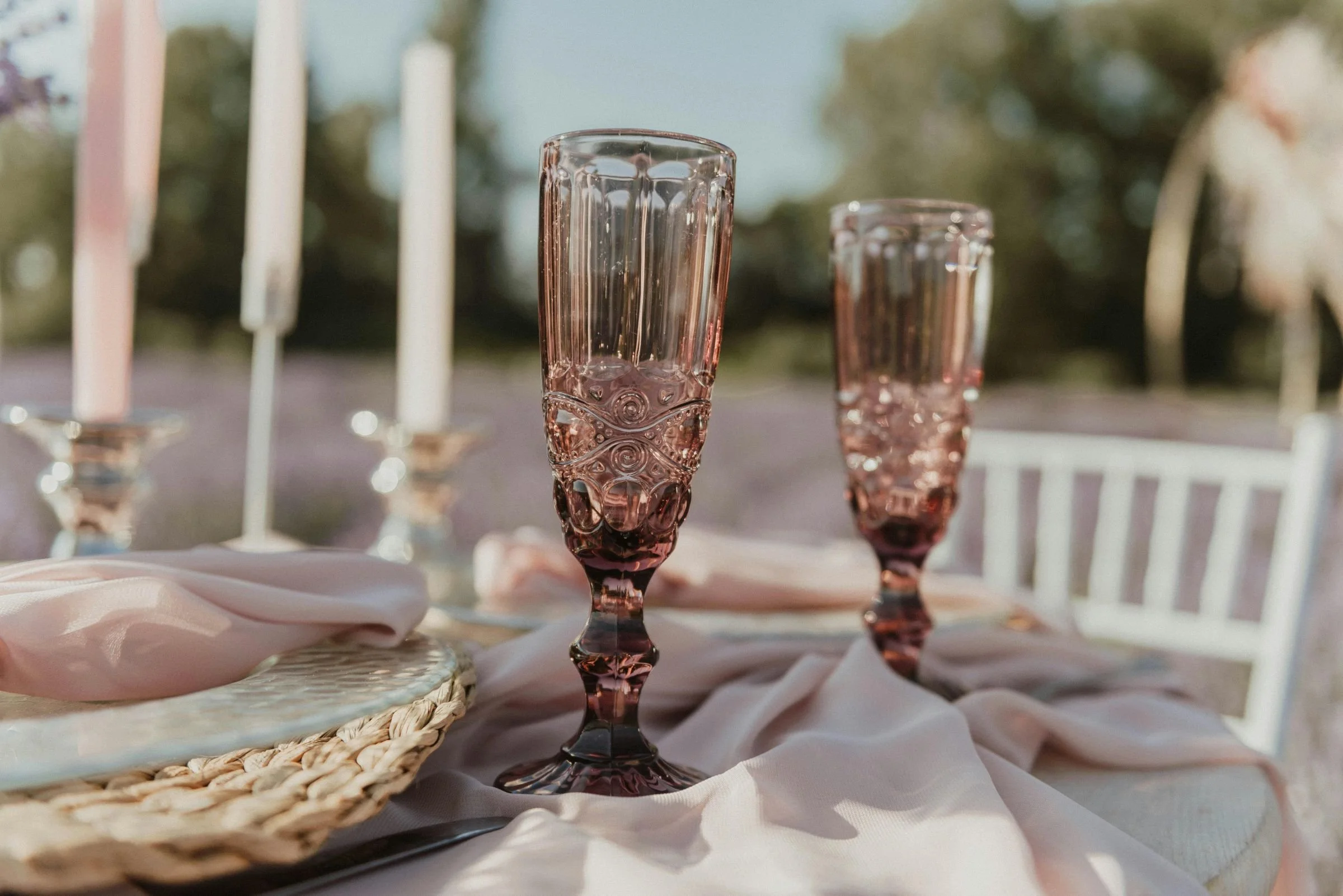 Two ornate pink glasses on a table set with soft pink fabric, candles, and woven placemats outdoors.
