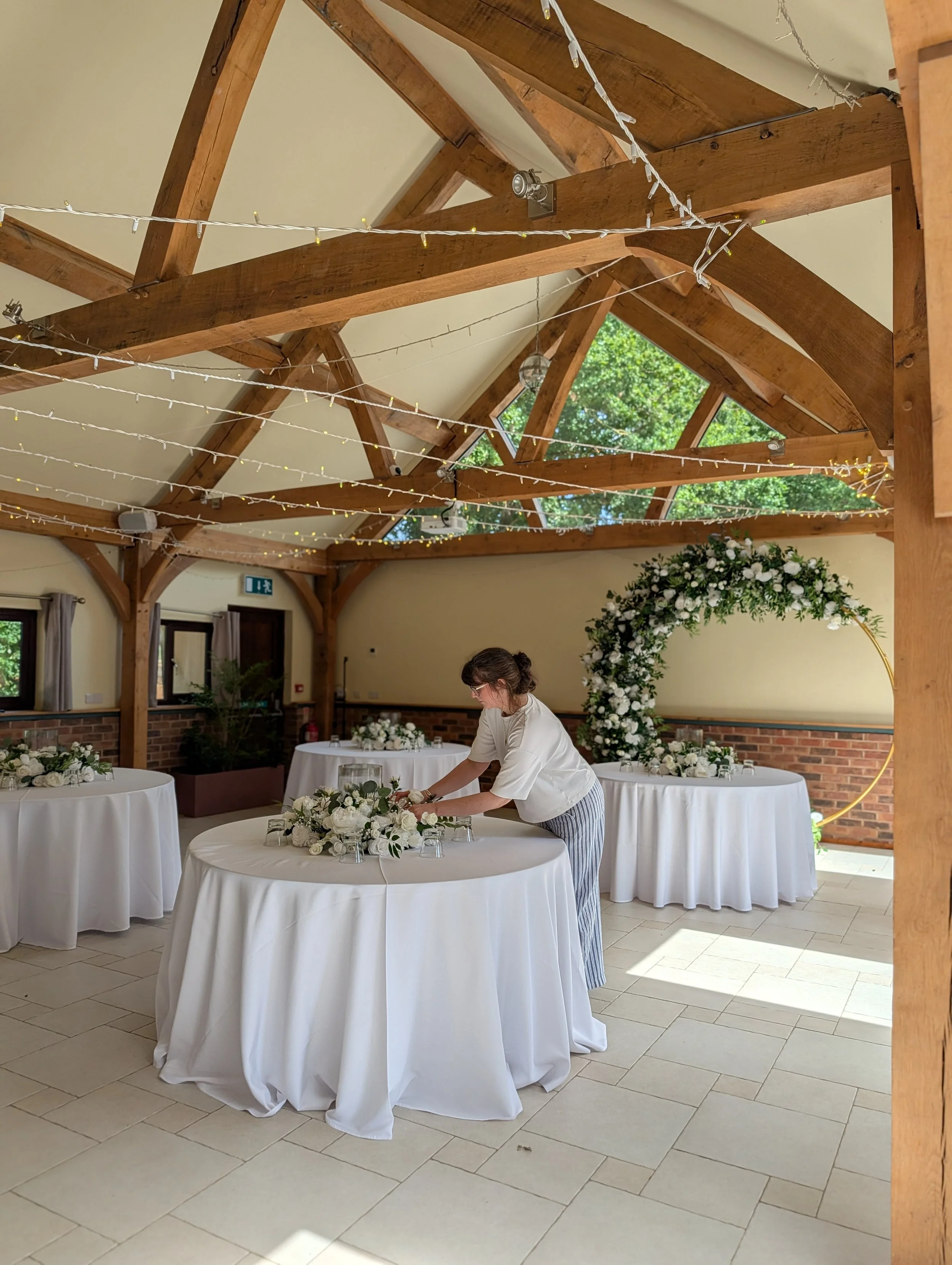An event stylist adjusts a floral centerpiece on a round table set for a wedding in a room with vaulted timber beams.