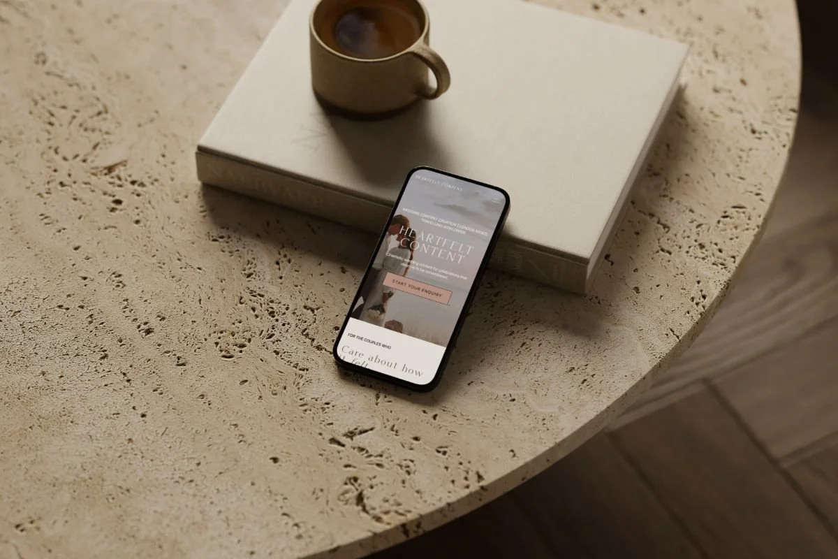 A mobile phone on a travertine table beside stacked books and a coffee cup, displaying the Heartfelt Content website homepage on a smaller screen