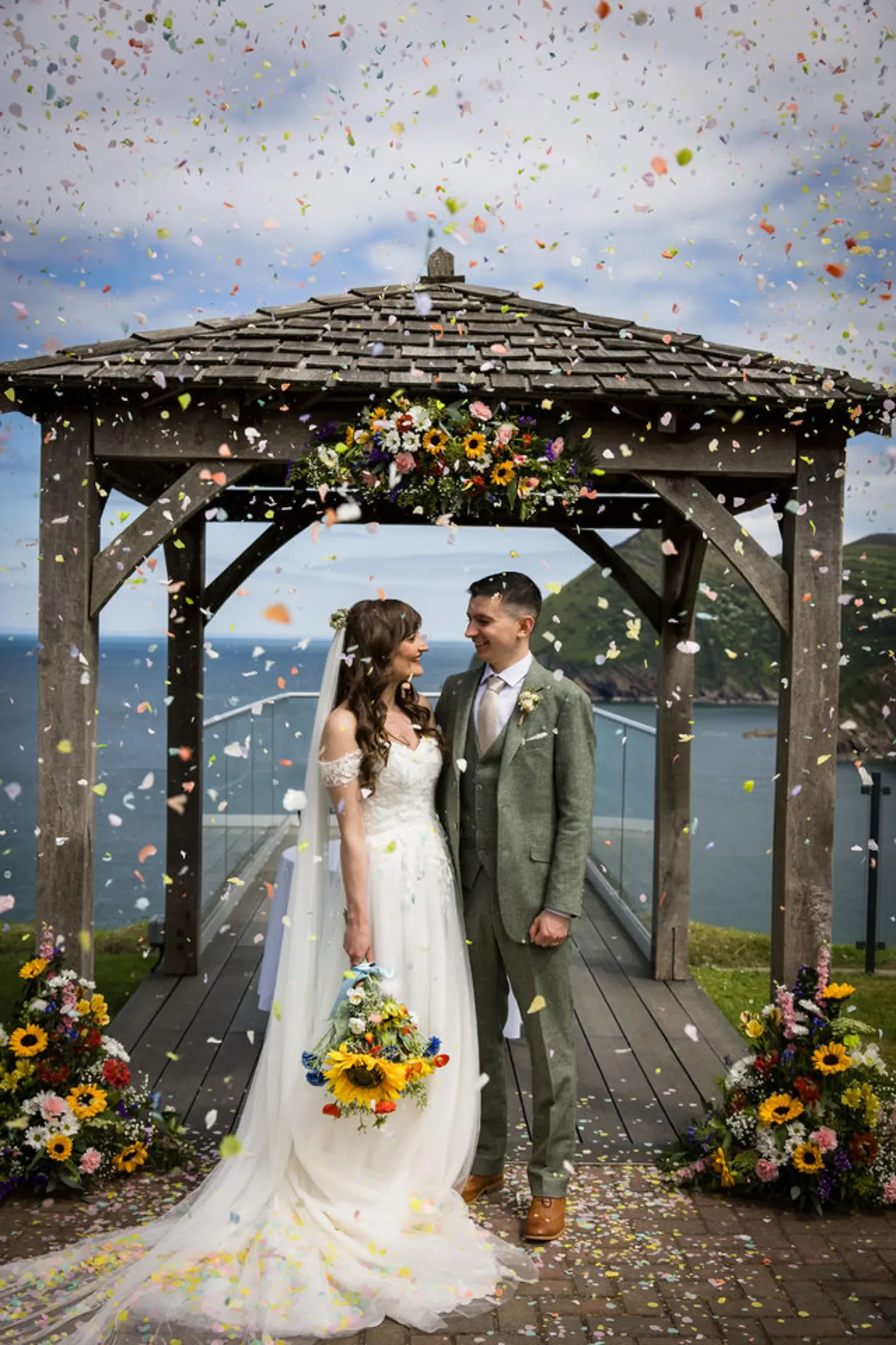 A bride and groom stand under a floral gazebo by the sea—perfect inspiration for wedding photographer branding with vibrant confetti joy.