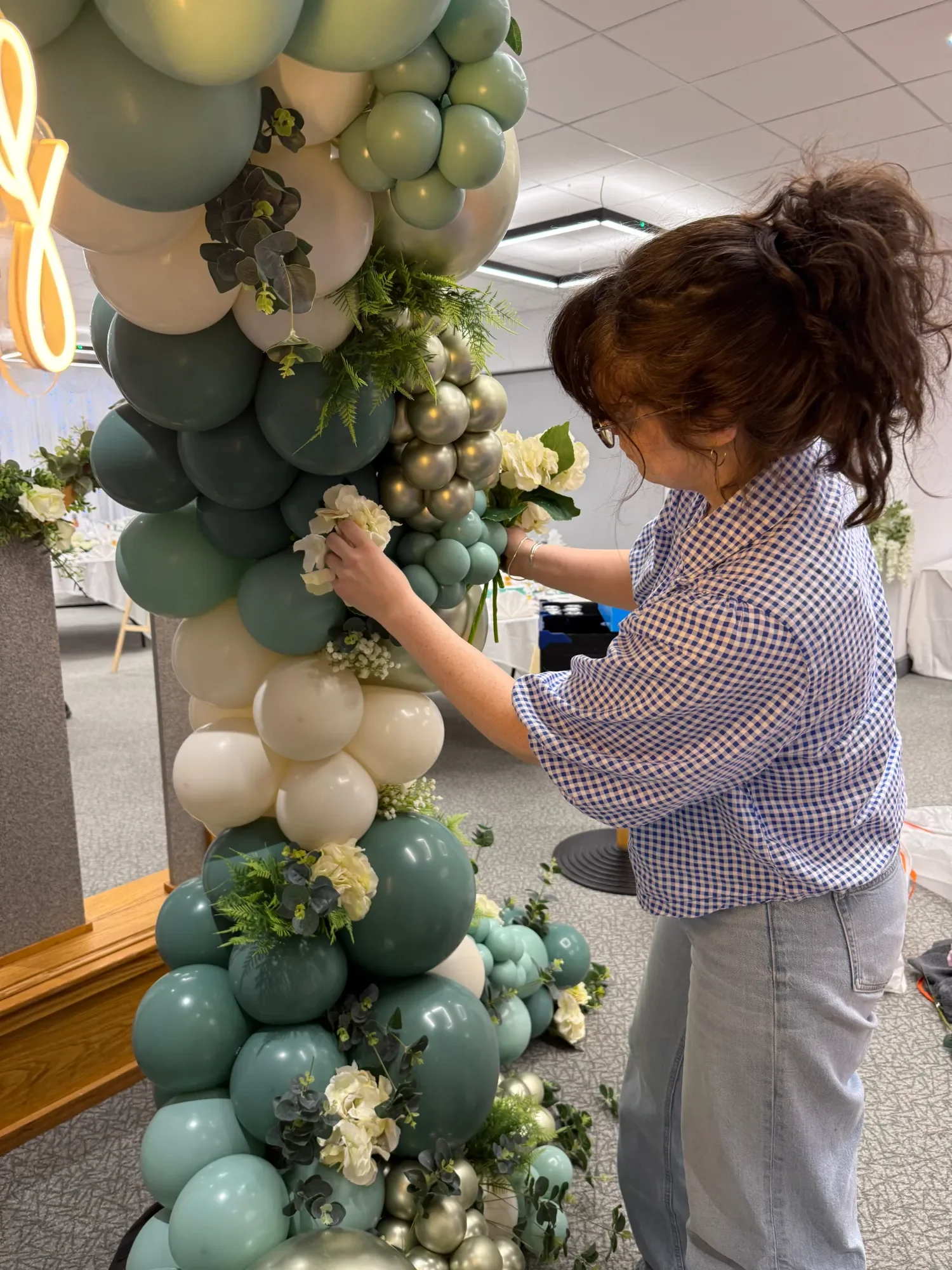 An event stylist in a blue gingham shirt arranging white flowers into a large balloon arch.