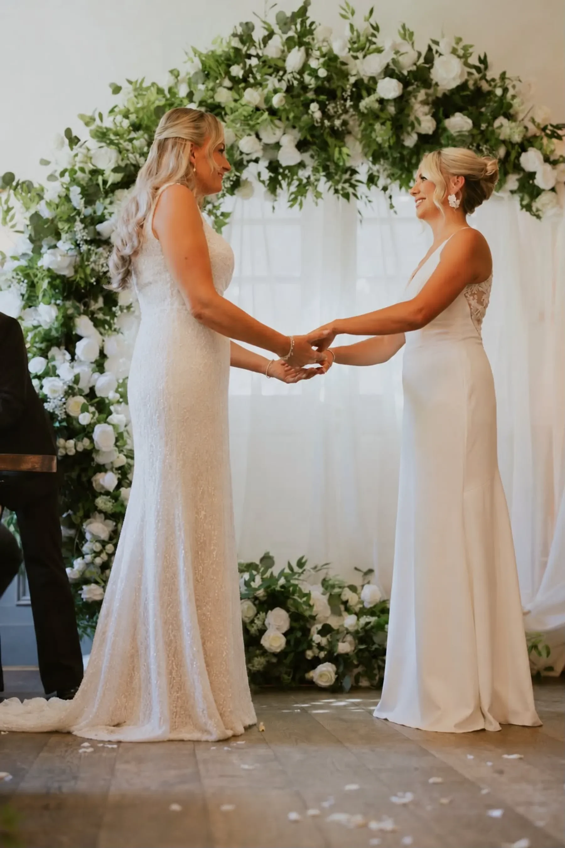 Two brides in white wedding dresses hold hands during a wedding ceremony in front of a large floral arch.