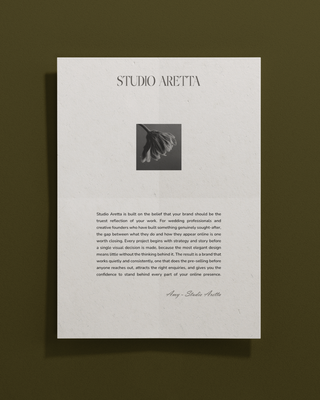 A white paper with black text titled 'STUDIO ARETTA', with a small black and white photo of a flower in the center, mounted on a green wall.