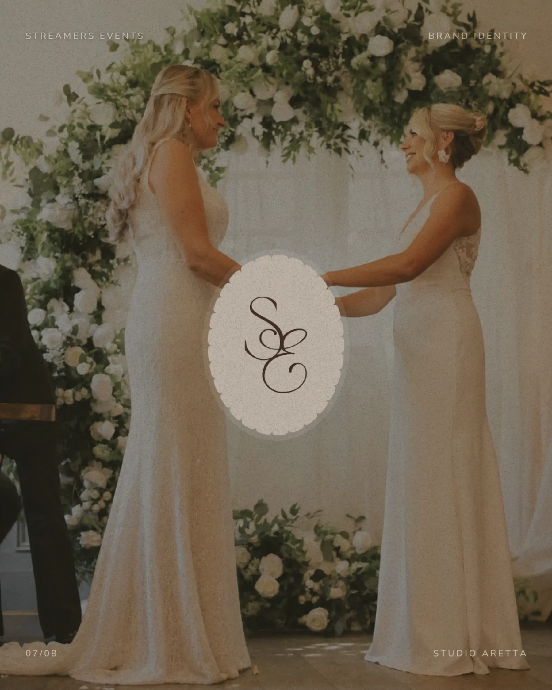 Two brides in white wedding dresses hold hands before a floral archway styled by an event stylist.