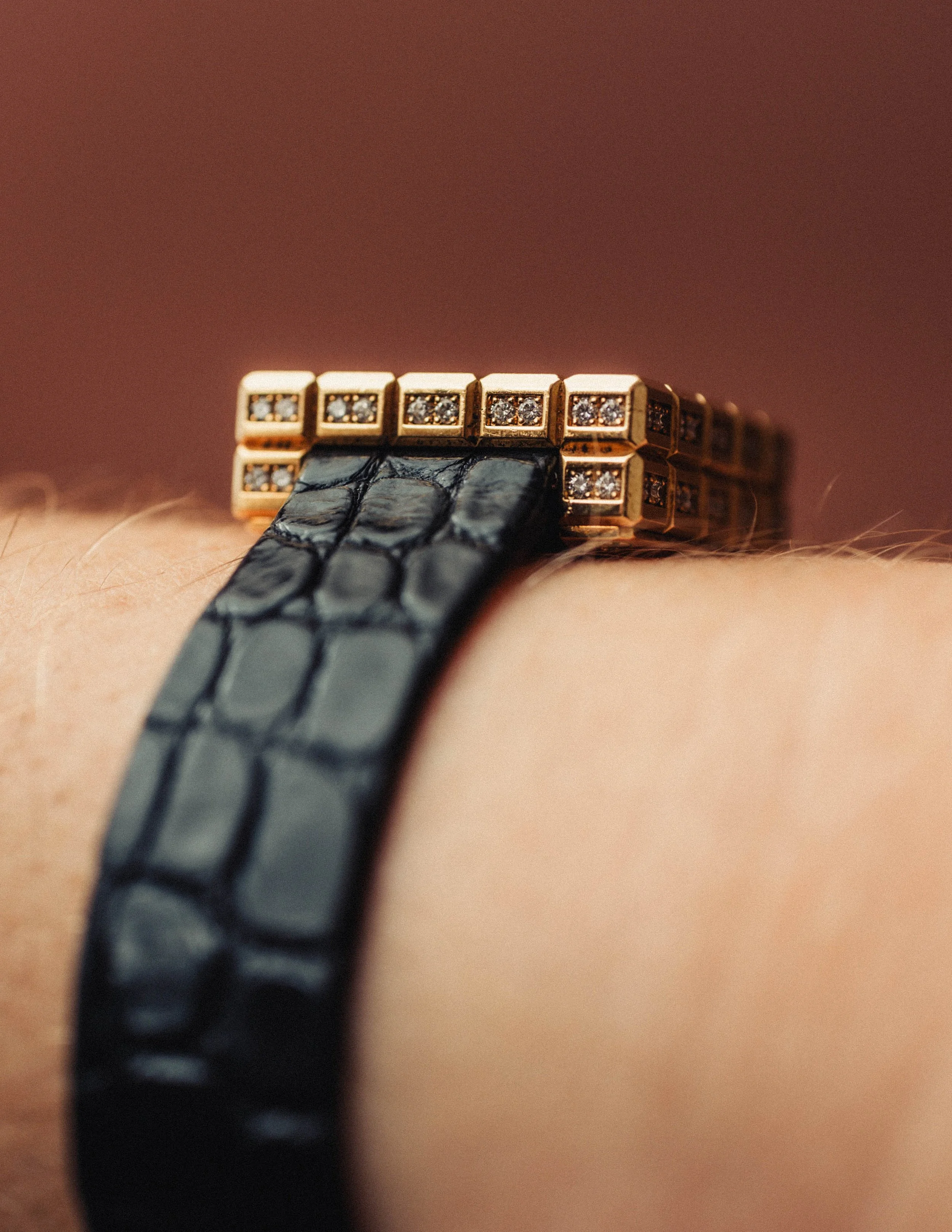 Close-up of a black leather watch strap with a gold buckle encrusted with small diamonds, on a person's wrist.