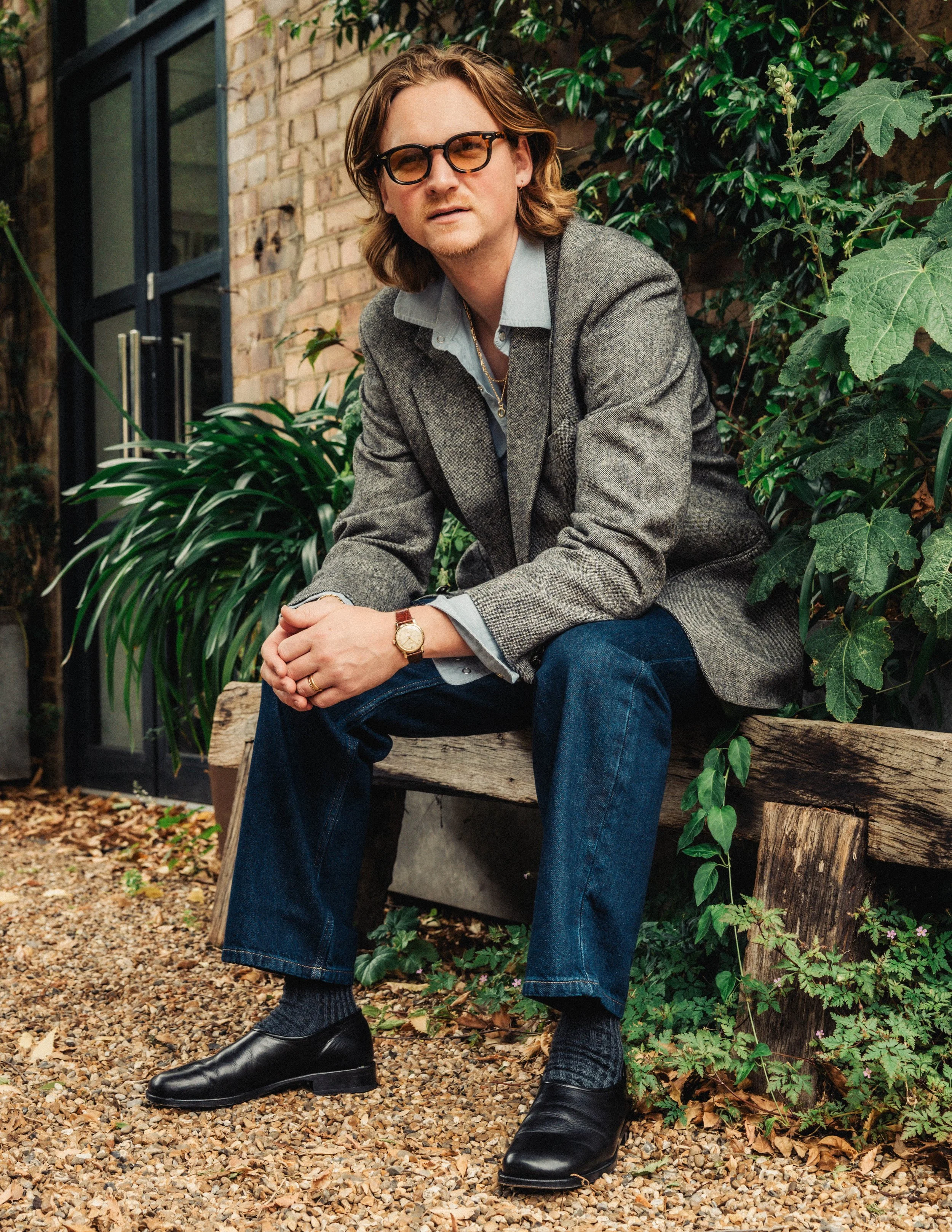 A man with medium-length wavy hair, wearing glasses, a gray blazer, a light blue shirt, dark blue jeans, and black shoes, sitting outdoors on a wooden bench surrounded by green plants and a brick building in the background.