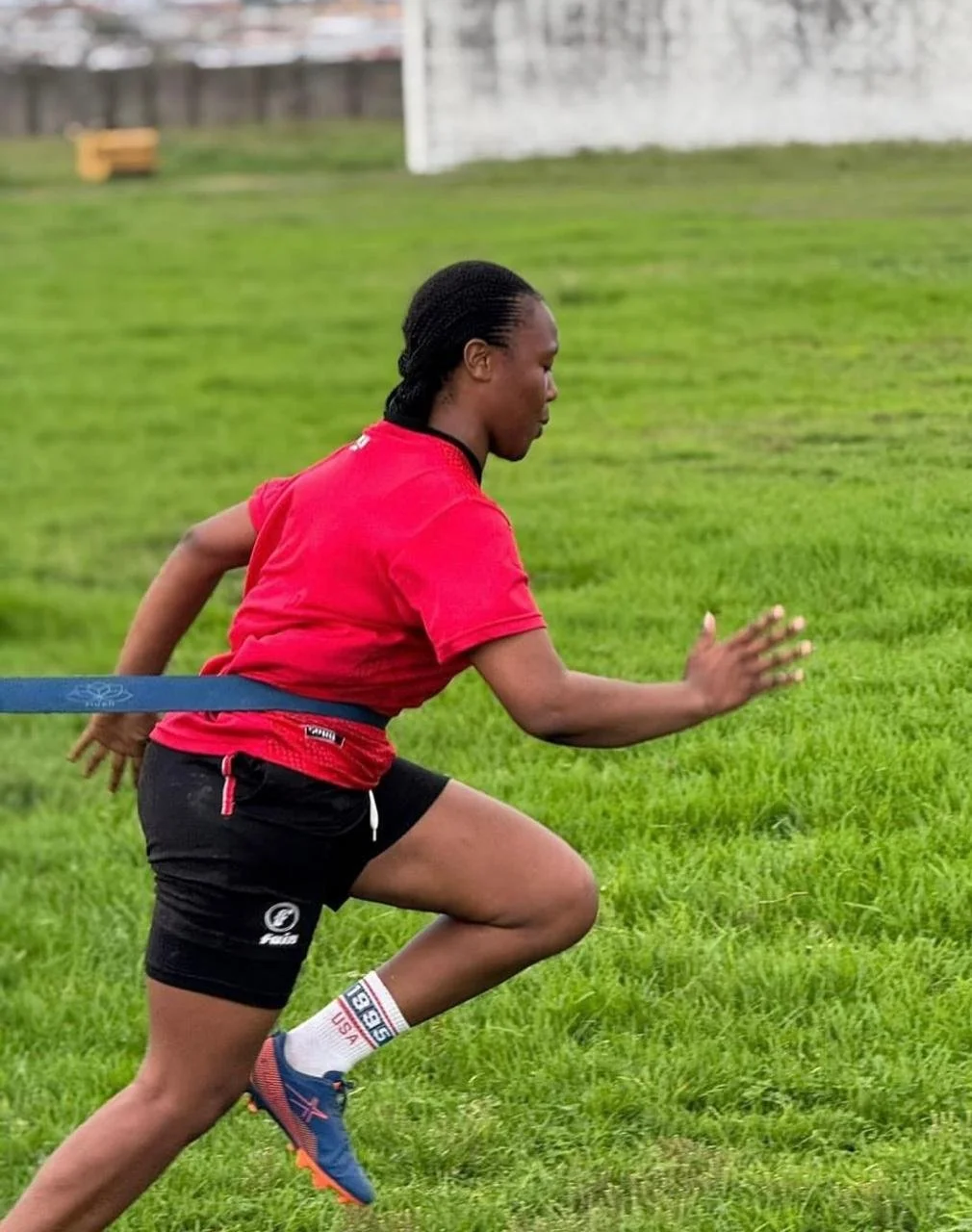 Three people standing together on a grassy field under a blue sky, with two women wearing sports uniforms and a man in casual athletic wear, smiling and posing for the photo.