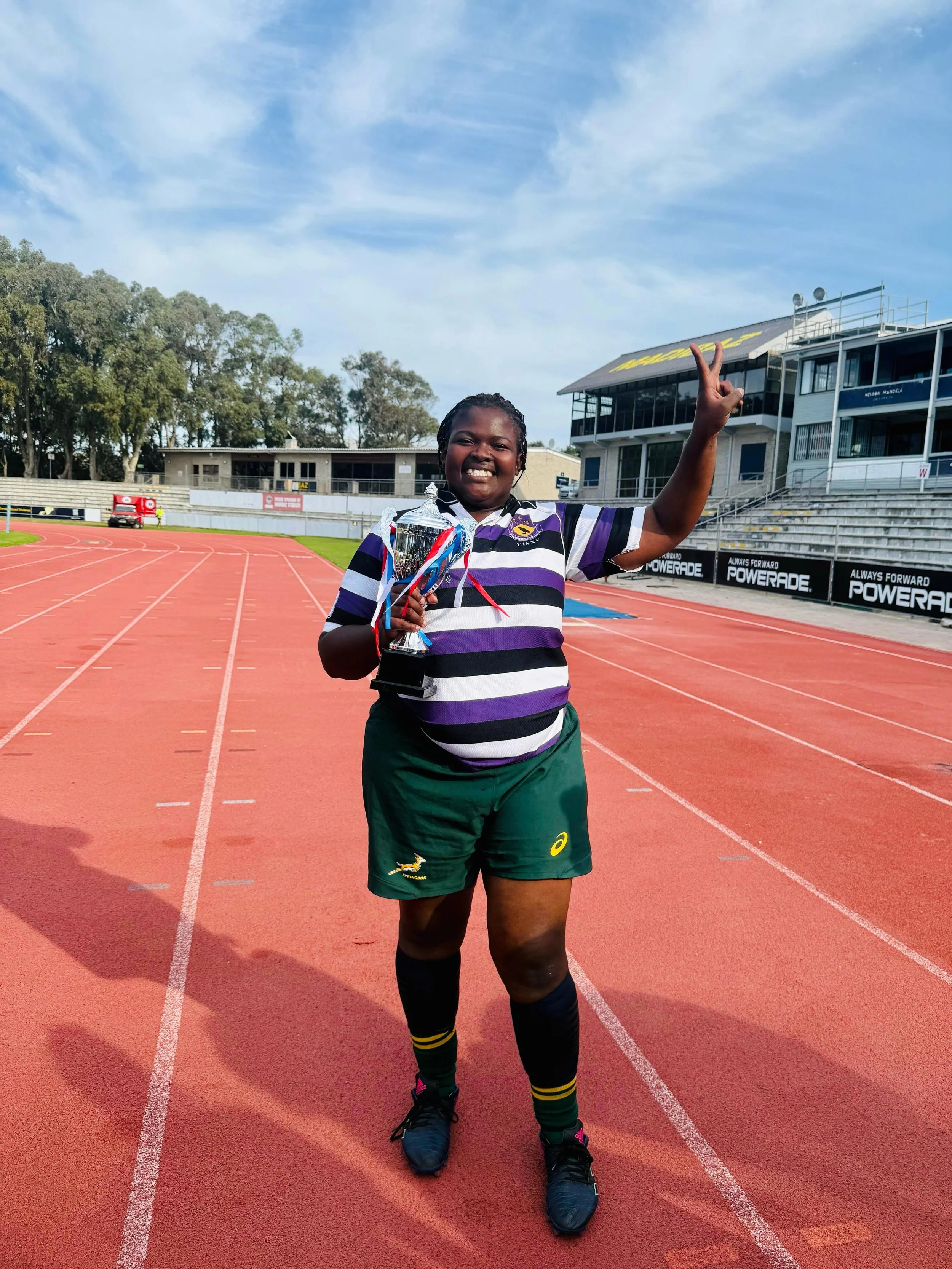 A young female athlete in a striped uniform holding a trophy, smiling and making a victory sign on a running track.
