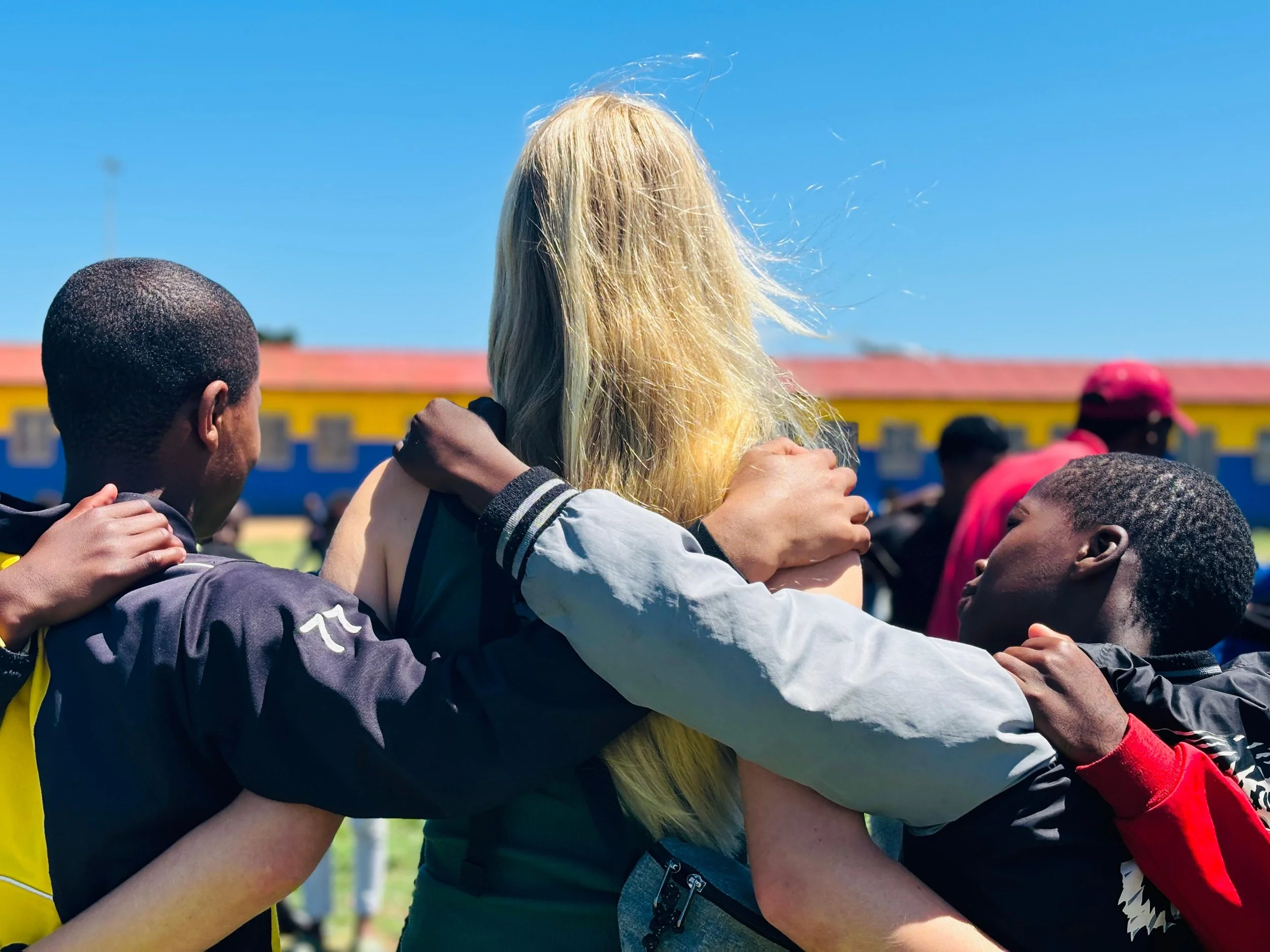 Two young men and a woman with long blonde hair in a huddle, outside on a sunny day, with a building and blue sky in the background.