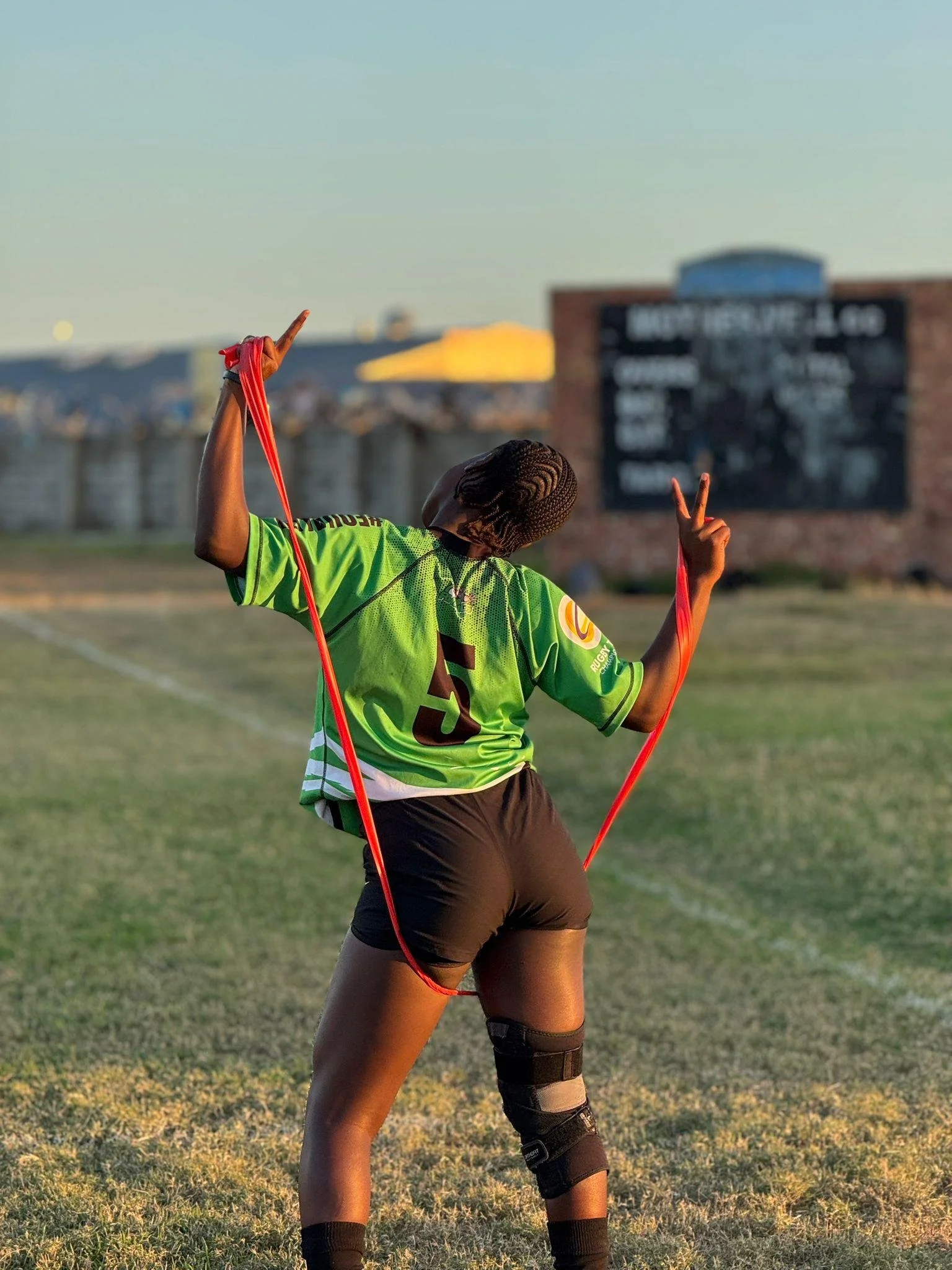A woman standing on a grassy field, wearing a green sports jersey and black shorts, with one knee brace. She is holding a red resistance band with each hand raised, making peace signs. Her hair is styled in braids, and she is facing away from the cam