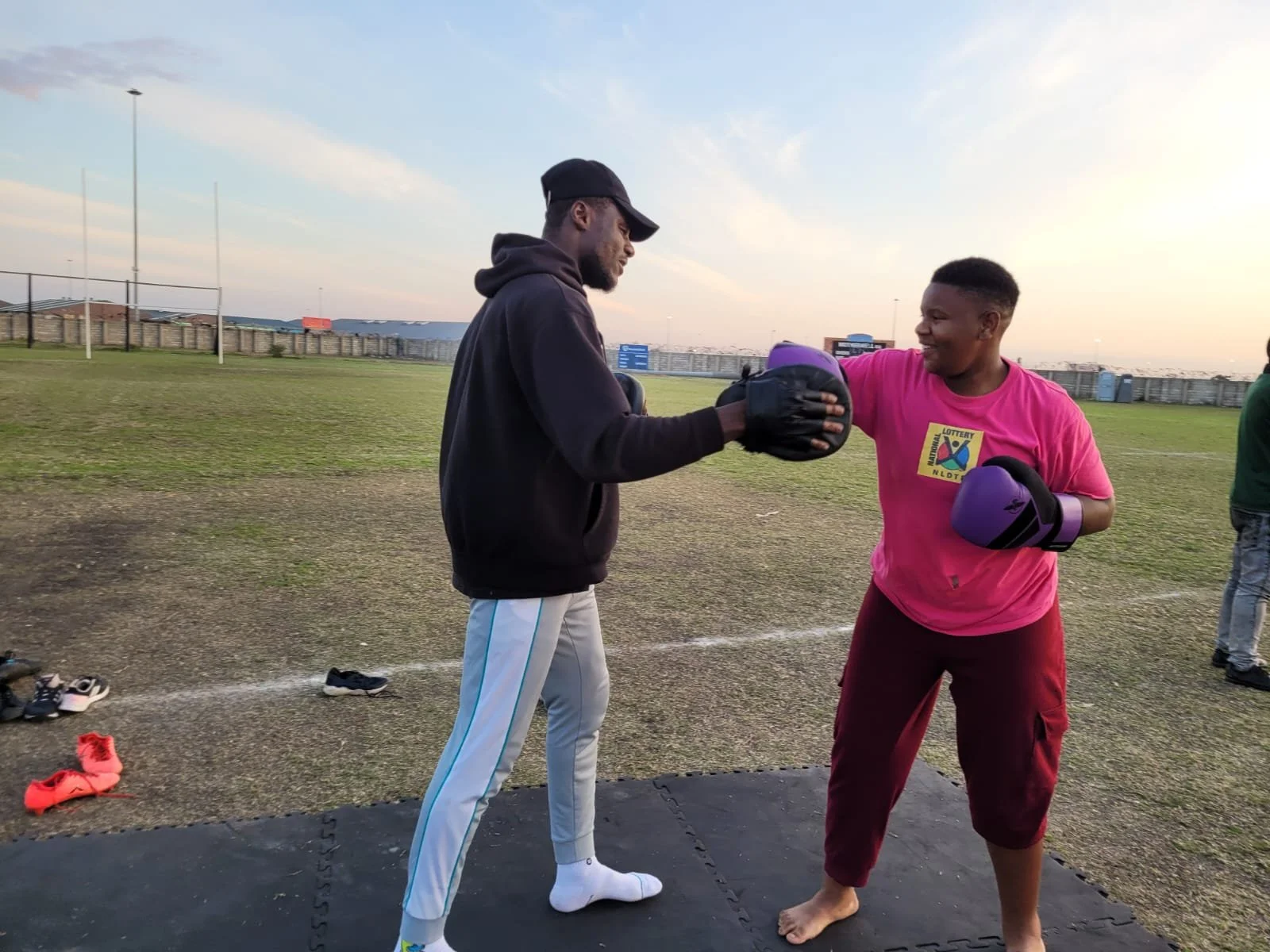 Two men practicing boxing outdoors, with one holding purple boxing gloves and protective gear, and the other wearing black gloves and a black hoodie, on a grassy field during sunset.