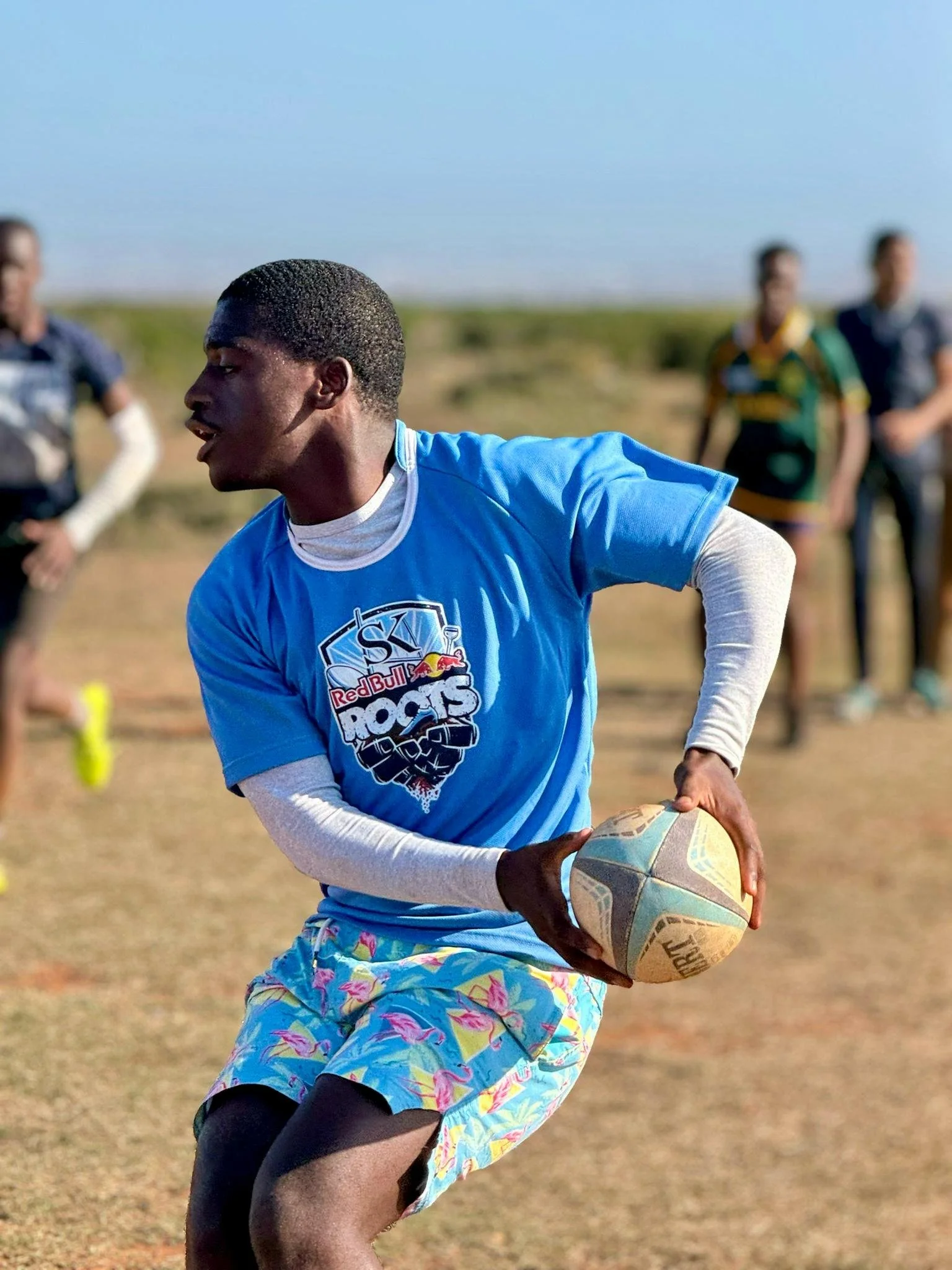 A young man holding a rugby ball on a field with a blue sky, wearing a blue Red Bull Red Bulls sports shirt and colorful shorts during a game.