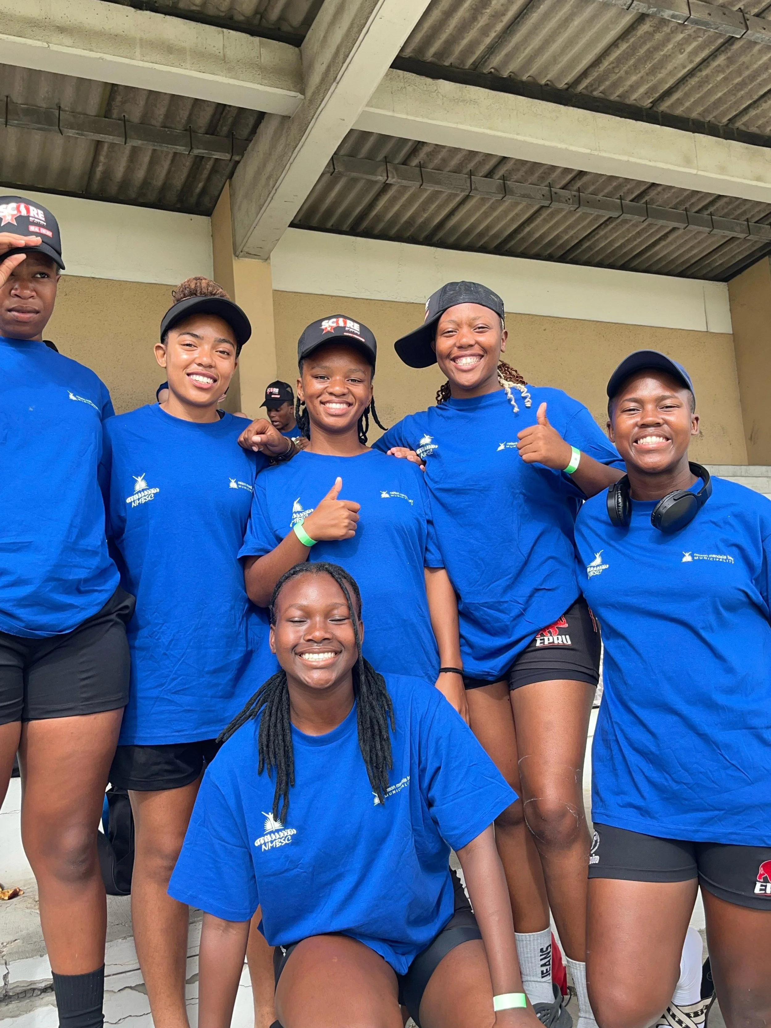 Group of young women in matching blue T-shirts and shorts posing together, smiling, with some giving thumbs up or wearing hats and headphones.