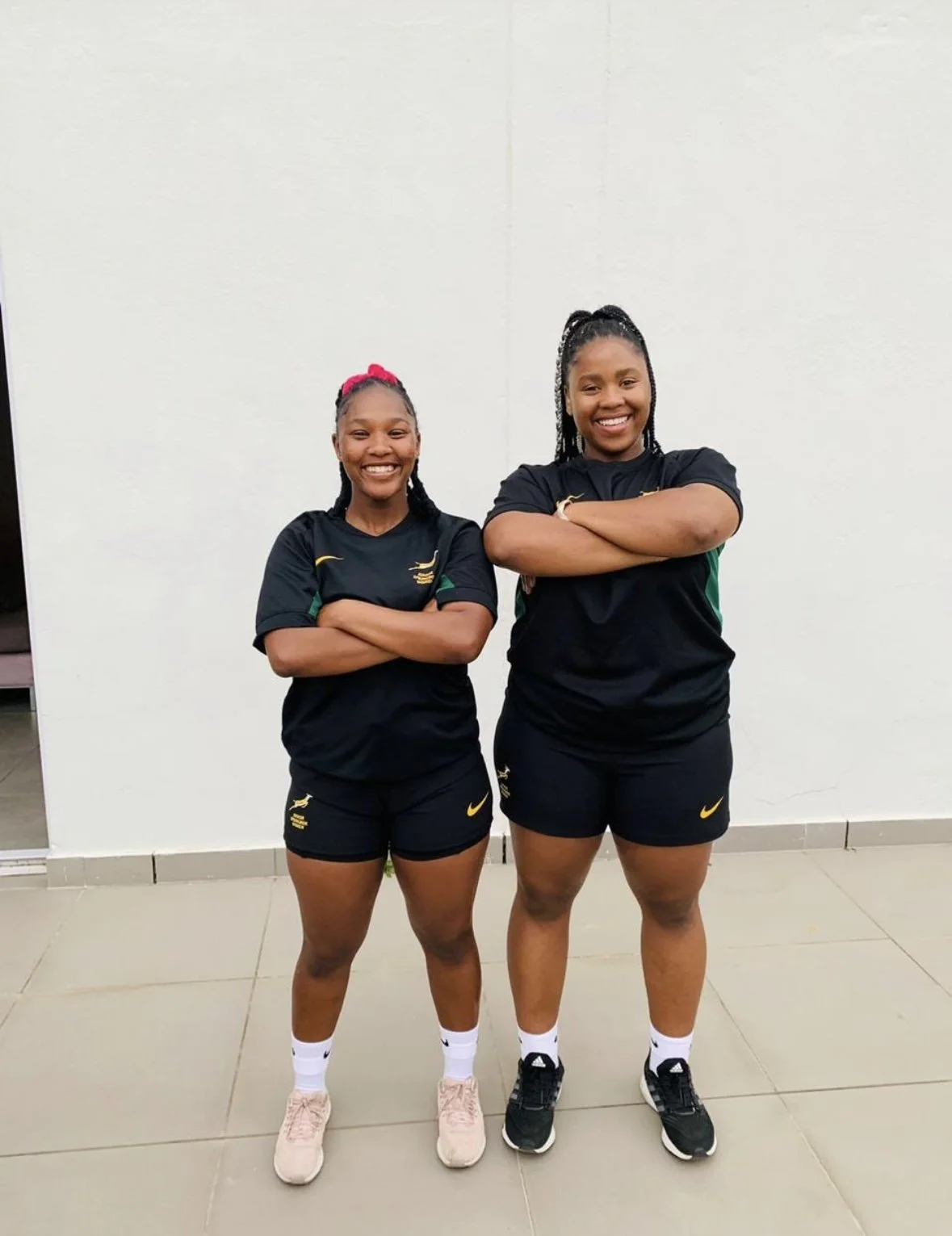 Two young women standing with arms crossed, wearing black athletic shirts and shorts, smiling at the camera, against a plain white wall.
